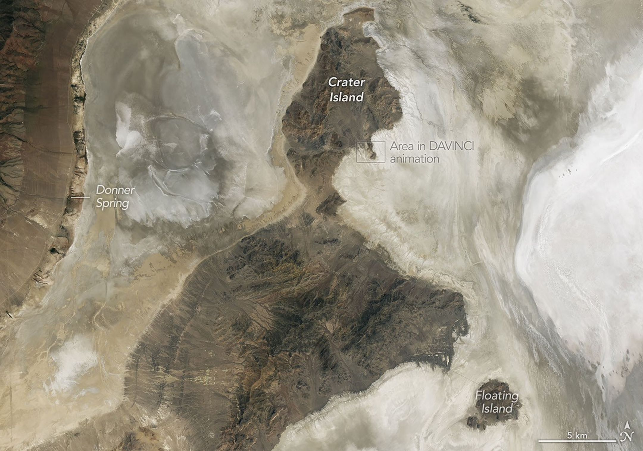 A nadir view shows Crater Island—a dark, linear mountain ridge in the center of the image—flanked by flat tan and white landscapes.