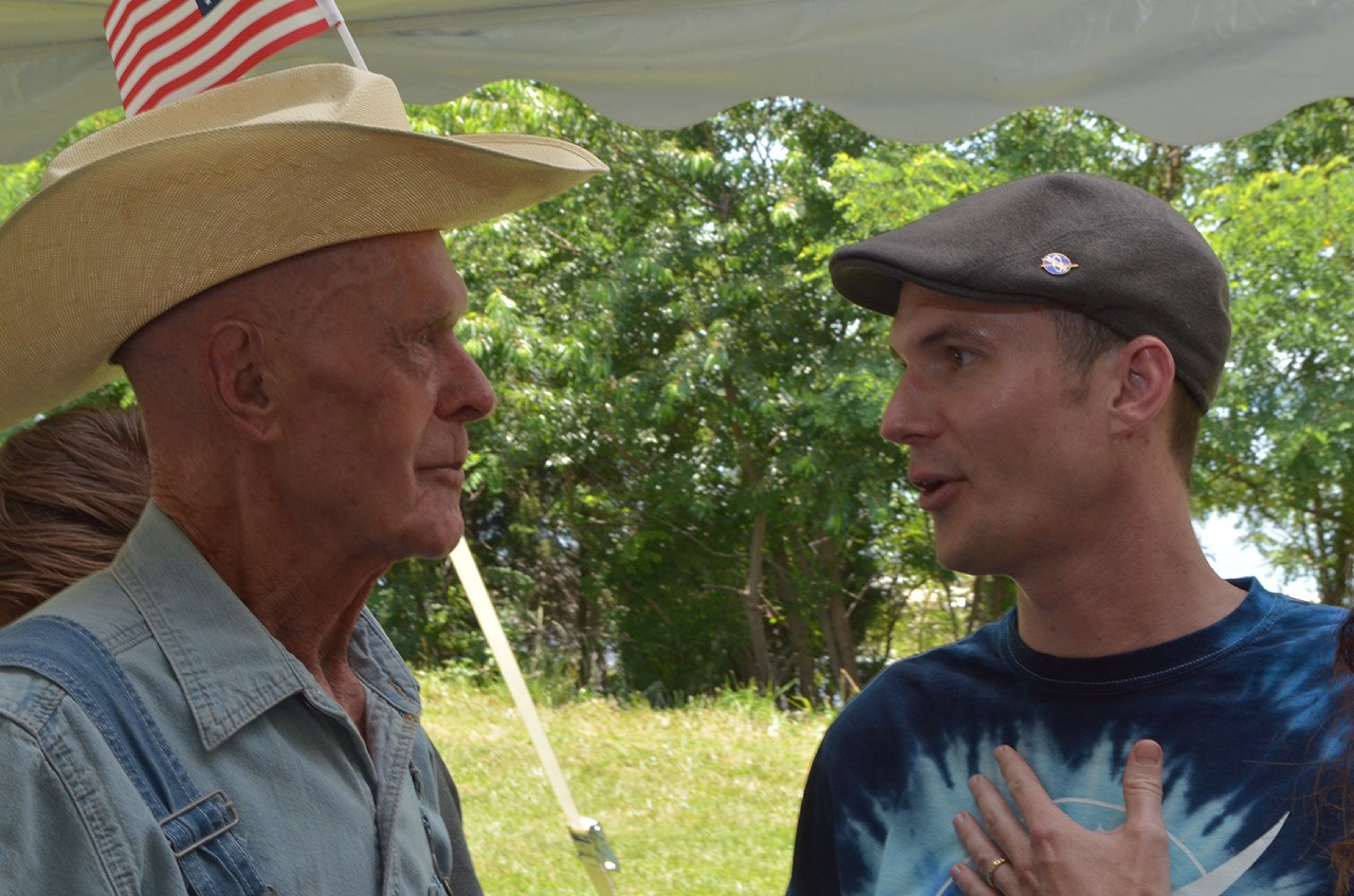 elder farmer on left in a straw hat talks to a man in a beret and tie-dye shirt in front of a nearby woodline