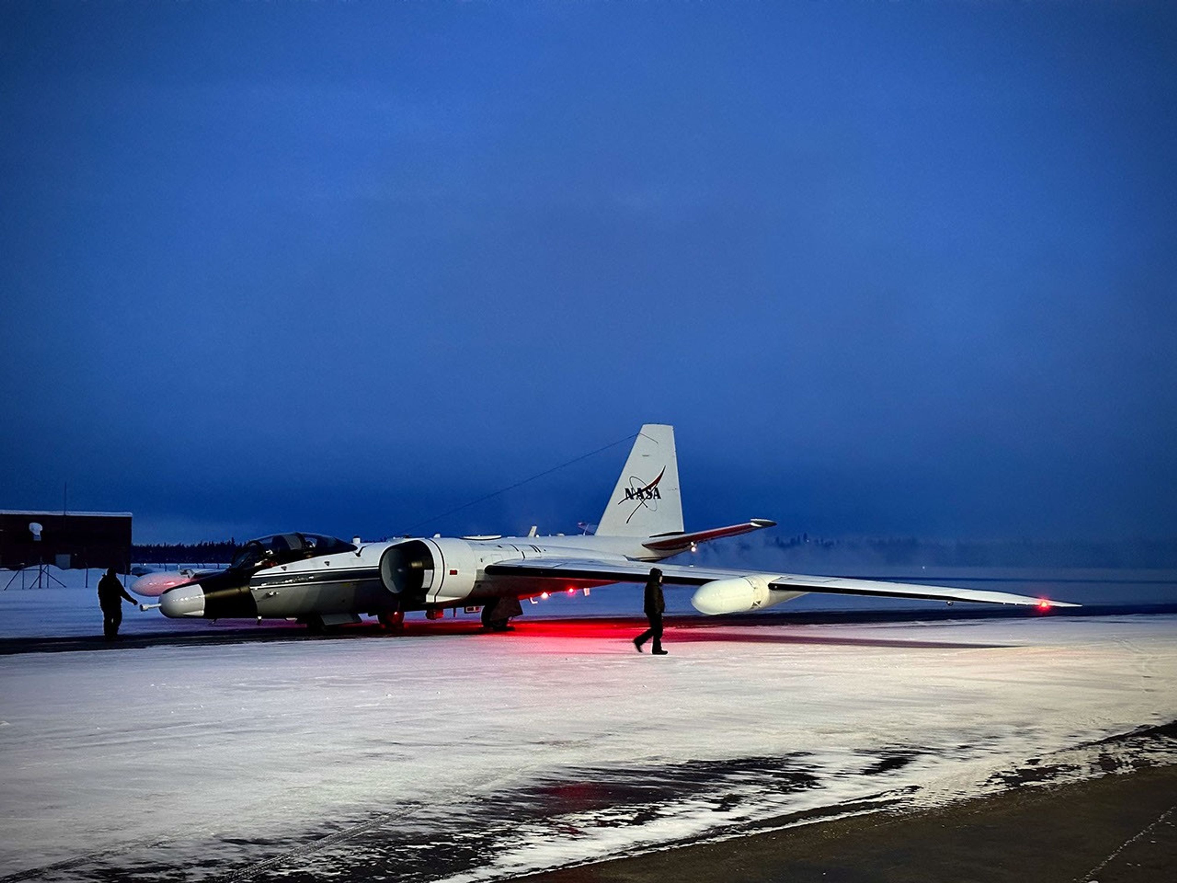 Two people walk in front of an airplane sitting on a snow covered runway as it prepares to study atmospheric particles over the Arctic.