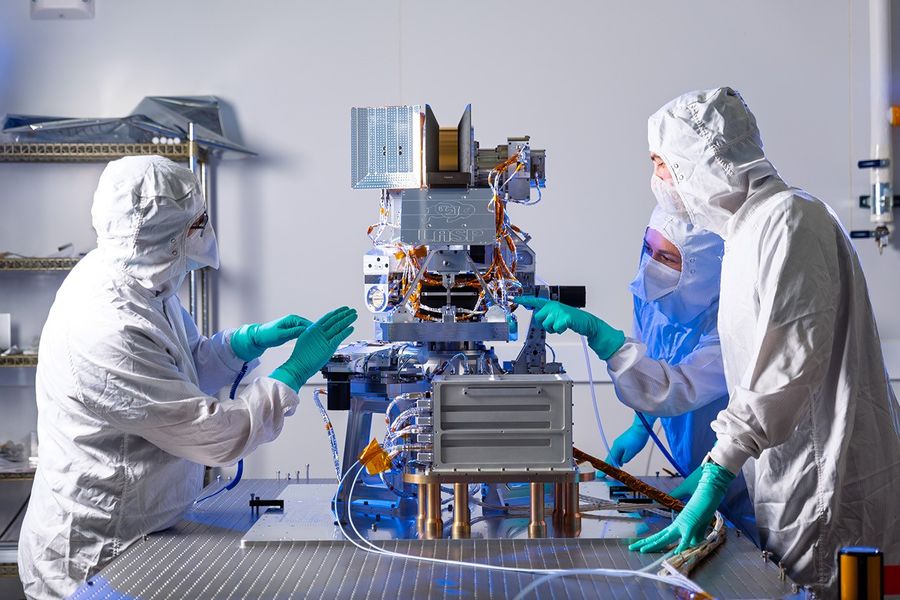 Three people in clean room suits stand around a piece of complex intrumnetation with wires and electronics exposed