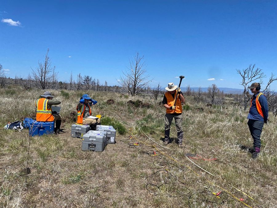 A person holds a sledgehammer while wearing an orange safety vest and standing in an arid landscape while other people look on.
