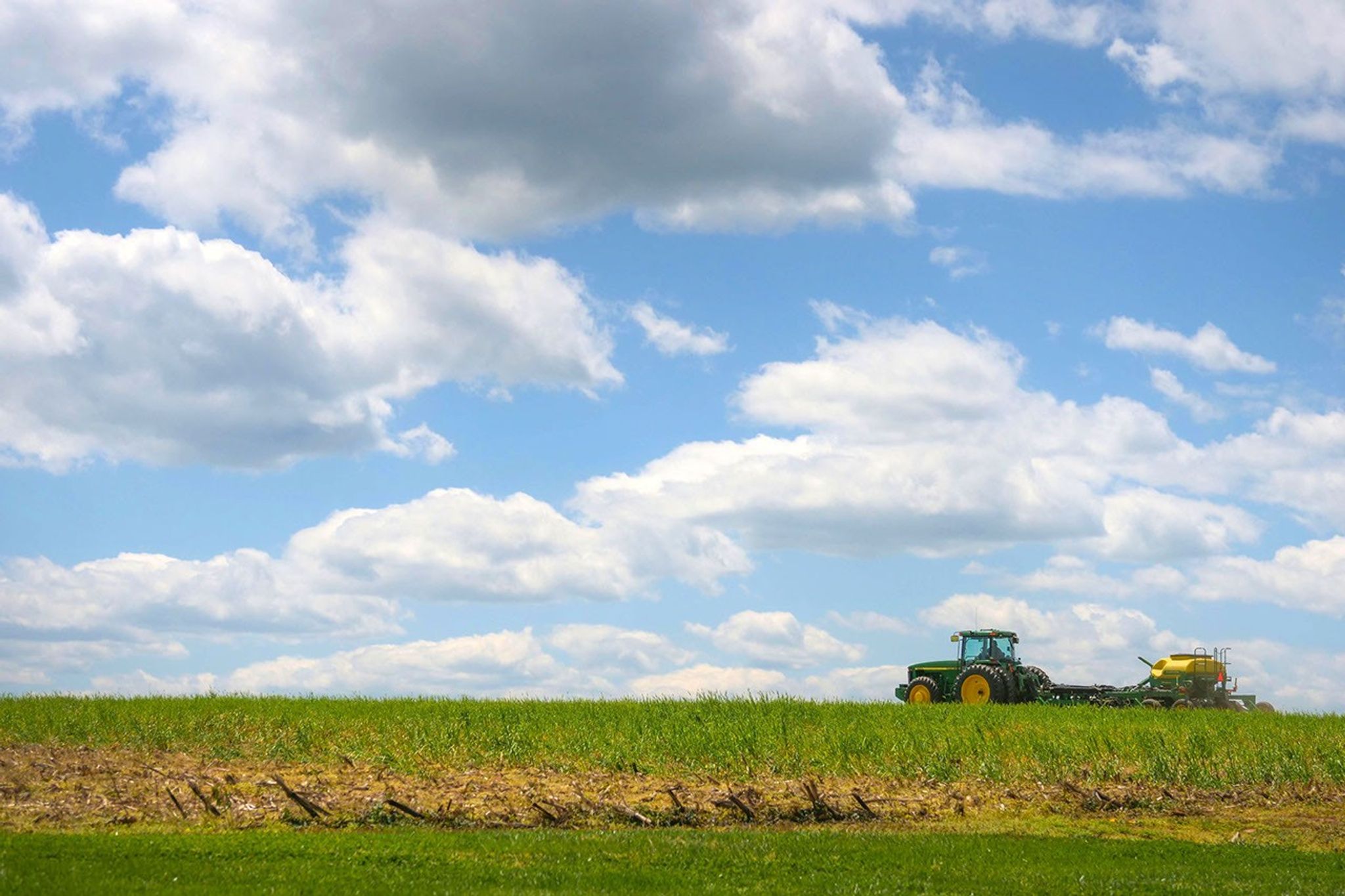 A green tractor travels through a field under a blue sky with scattered clouds.