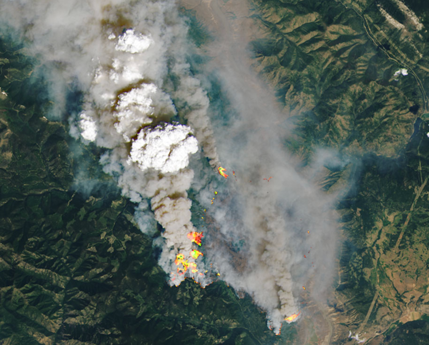 A white cloud as viewed from above rises over gray smoke streaming from fires in a mountainous region as seen from space