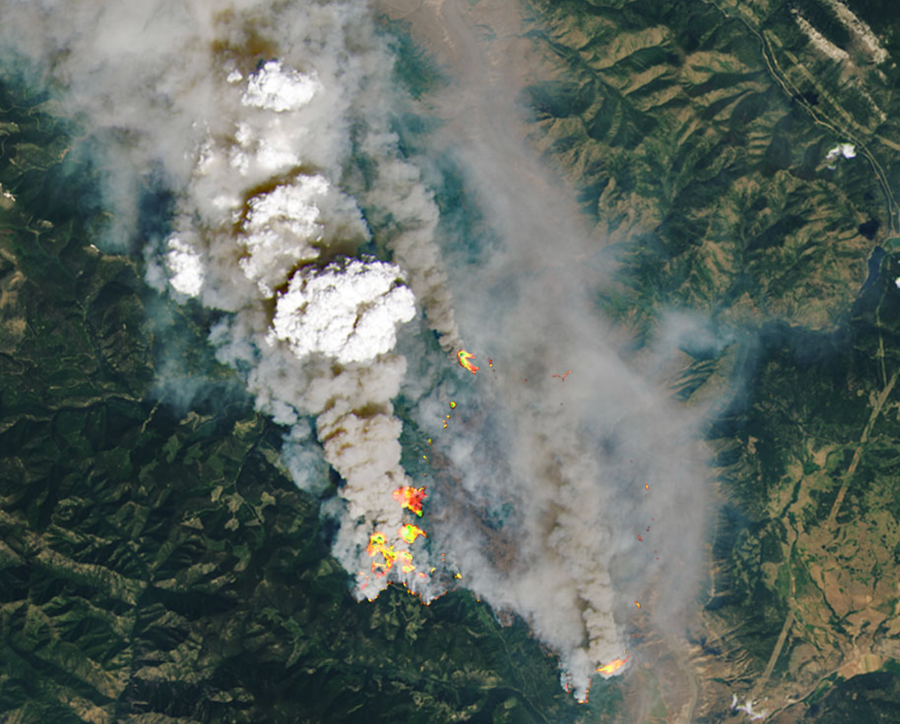 A white cloud as viewed from above rises over gray smoke streaming from fires in a mountainous region as seen from space