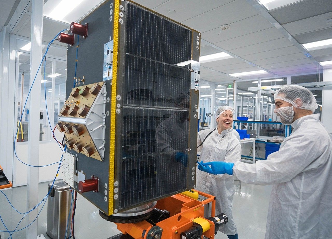 Two people stand next to a large box shaped device on a pedastal. The people are in a room surrounded by floor to ceiling glass walls and wearing white cover coats and hairnets. The person on the right is also wearing a covering over their facial hair.