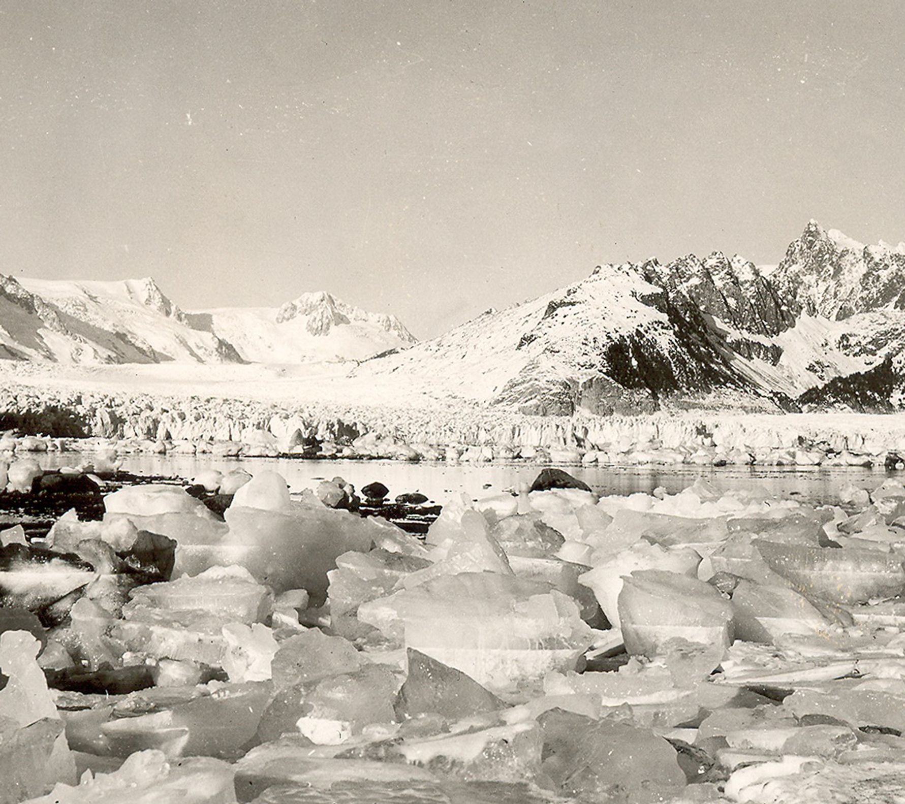 Looking north from the west shoreline of Harris Bay, Kenai Fjords National Park, Kenai Mountains, Alaska. The rocky shoreline in the foreground is covered by small icebergs, calved by the retreating Northwestern Glacier. During the roughly 60 to 80 years between photographs, Northwestern Glacier retreated some 6 miles (10 kilometers) to the northwest, out of the field of view.  U.S. Geological Survey, Department of the Interior. Earlier image is from a postcard, photographer unknown, courtesy of Kenai Fjords National Park. Later image is a USGS photograph by Bruce F. Molina.