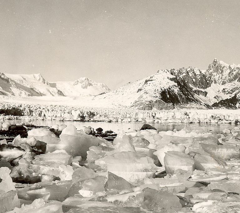 Looking north from the west shoreline of Harris Bay, Kenai Fjords National Park, Kenai Mountains, Alaska. The rocky shoreline in the foreground is covered by small icebergs, calved by the retreating Northwestern Glacier. During the roughly 60 to 80 years between photographs, Northwestern Glacier retreated some 6 miles (10 kilometers) to the northwest, out of the field of view.  U.S. Geological Survey, Department of the Interior. Earlier image is from a postcard, photographer unknown, courtesy of Kenai Fjords National Park. Later image is a USGS photograph by Bruce F. Molina.