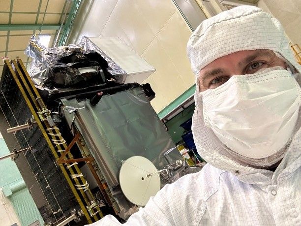 A man stands on the right side of the image seen from the shoulders up. He is wearing a white clean suite that covers over his head and forehead as well as a white mask that covers his nose and mouth. Behind him and to the left in the image is a spacecraft, PACE. Several parts of the spacecraft are covered in a silver-colored foil-like material. The room that the man and the spacecraft are in has very large ceilings which are seen in the background of the selfie.