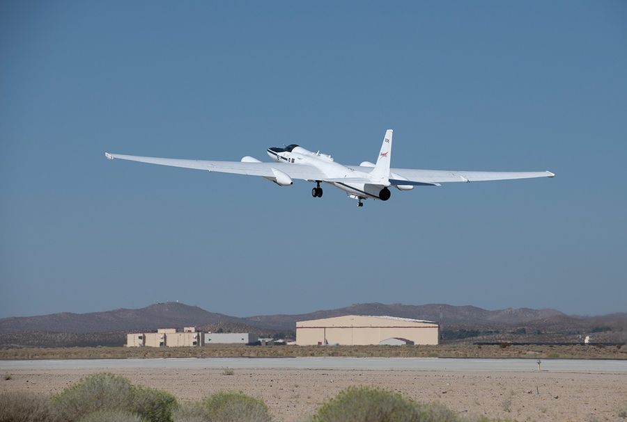 A plane takes off from a runway, seen mostly from the rear of the plane. It is pointed upwards and flying from the right to the left of the image. It is centered in the image. The horizon lies about a quarter of the way up the image and above the horizon are bright blue skies.