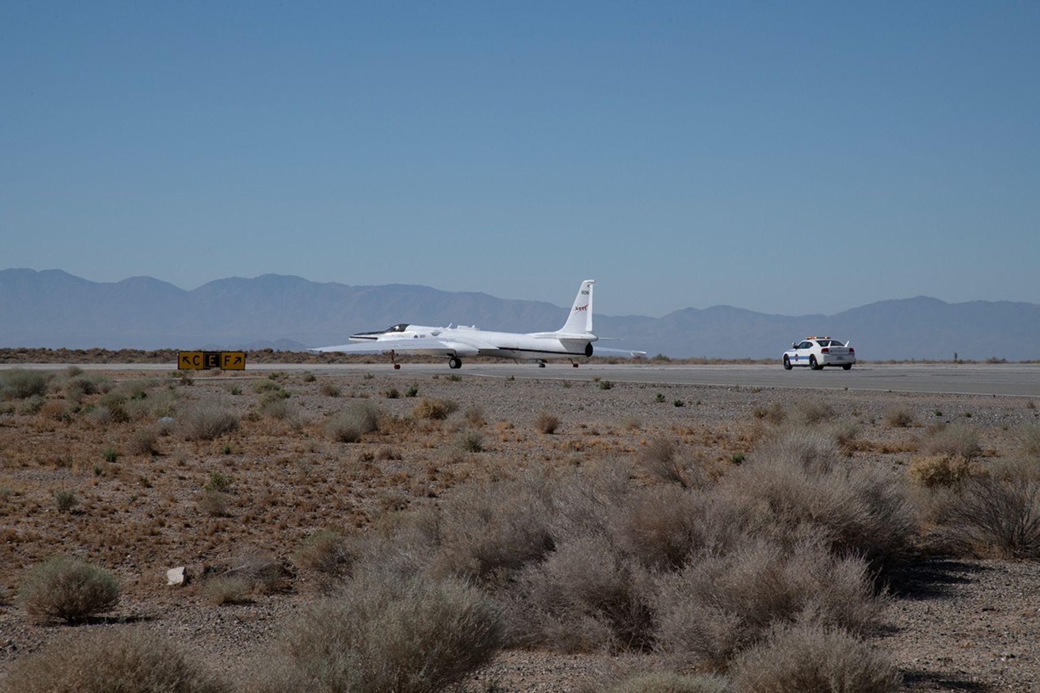 A plane rests on a runway, centered in the image. A car follows the plane on the runway. In the foreground is a desert landscape and the background shows a mountain range hidden by haze and bright blue skies.