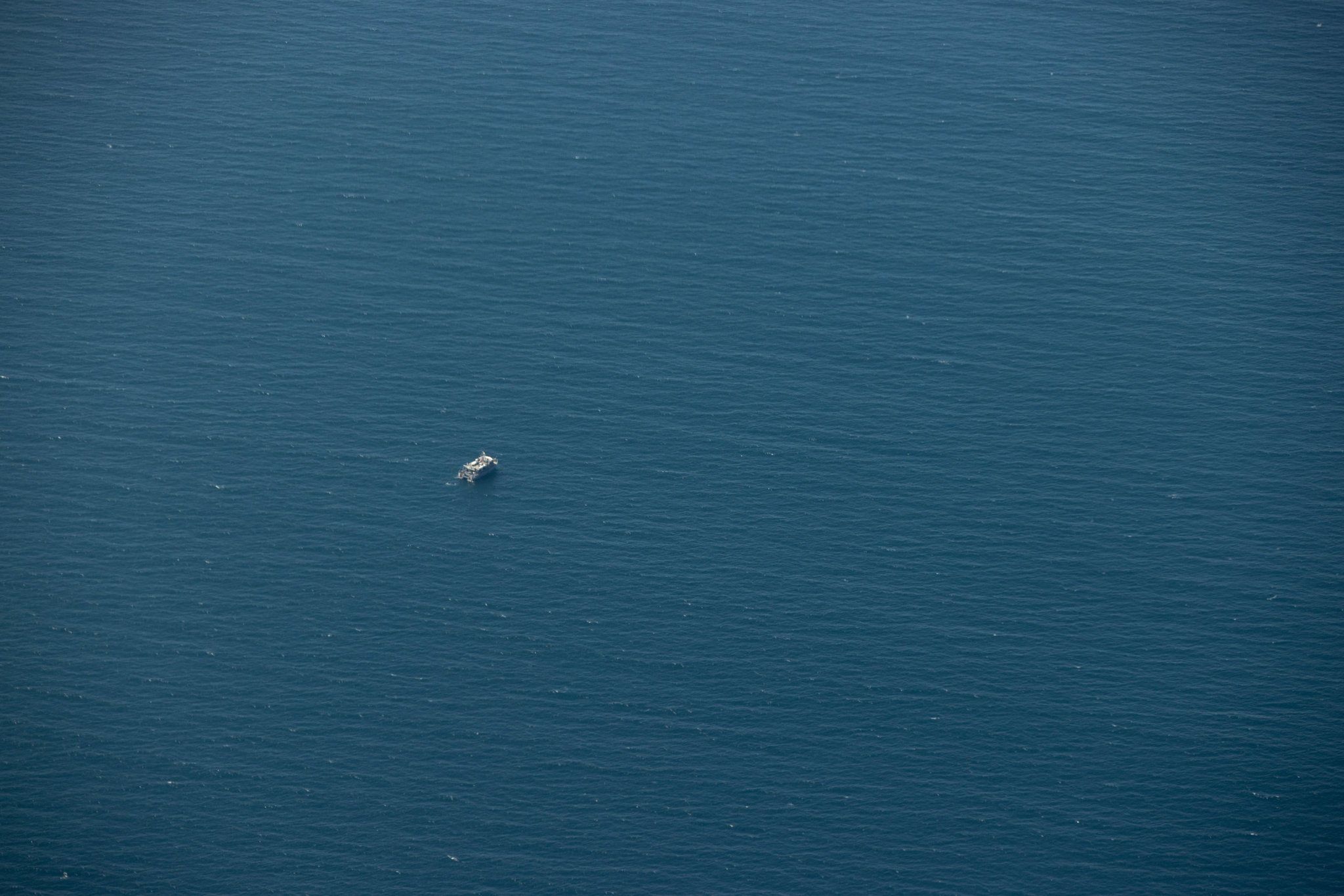 The image is primarily a deep blue of the ocean, with small ripples of waves seen. It is taken from high above the ocean. There is a small white dot in the center left of the image, which is the Shearwater research vessel as seen from the sky.