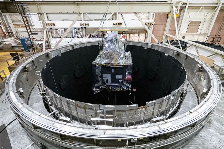 Centered in the screen is the observatory, covered in a reflective foil-like material. The observatory is being lowered into a dark, hollow, circular chamber, the TVAC testing chamber.
