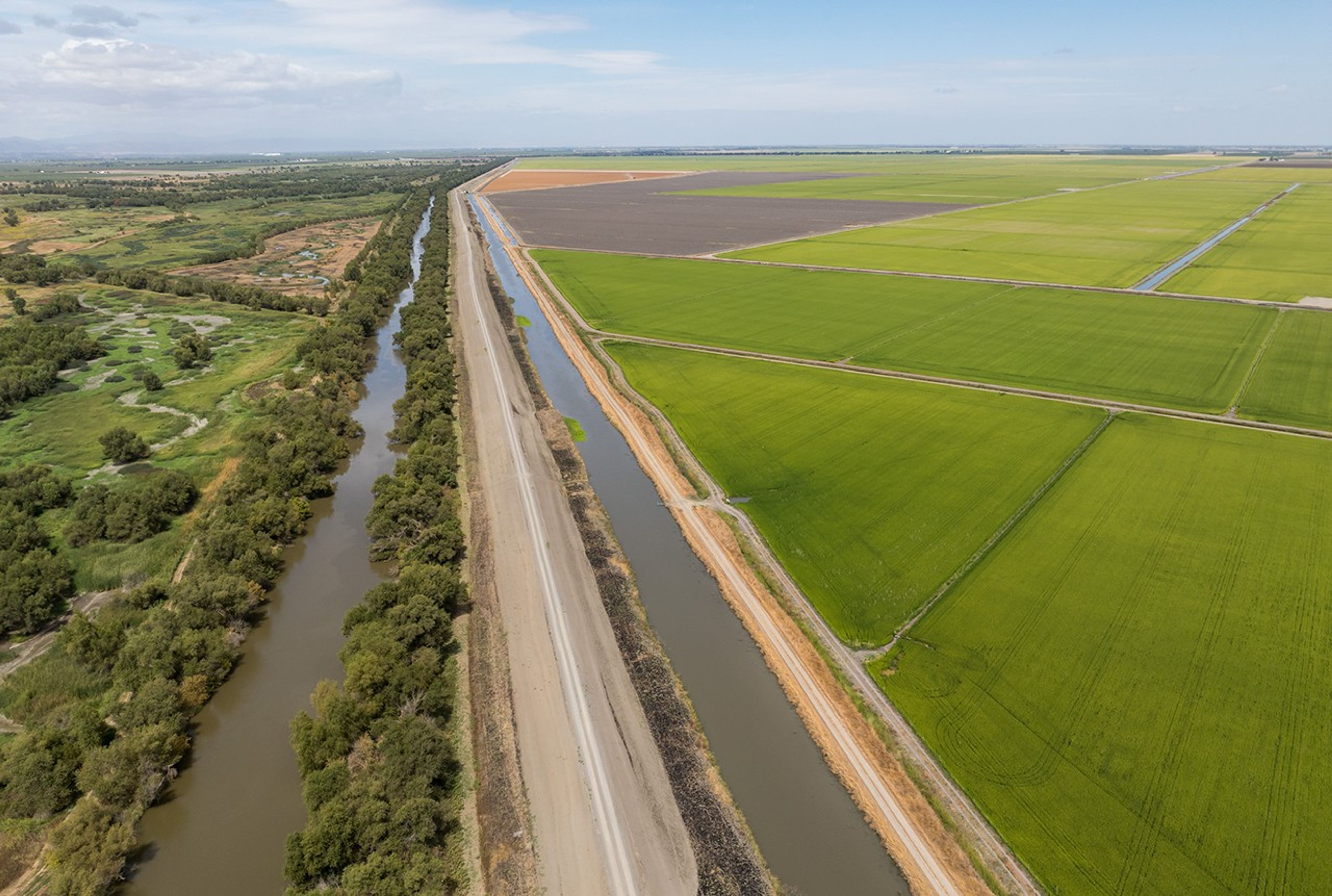 Aerial photo of California agricultural fields with irrigation canals and tree-lined waterways under a partly cloudy sky.