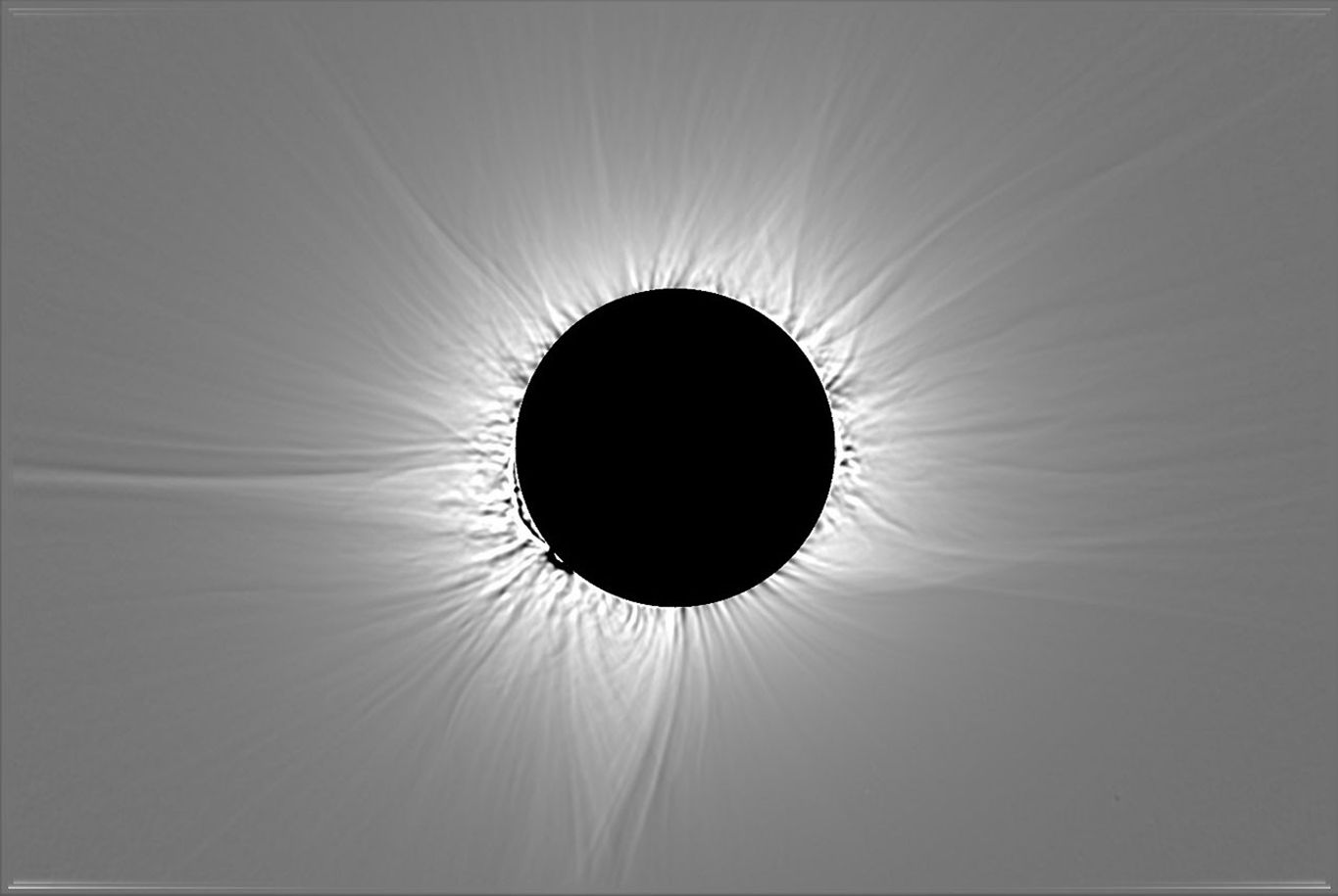 An image of a total solar eclipse shows the Moon as a black disk at the center surrounded by white rays extending outward around the Moon, set against a gray background. Some of the rays are longer or thicker than others.