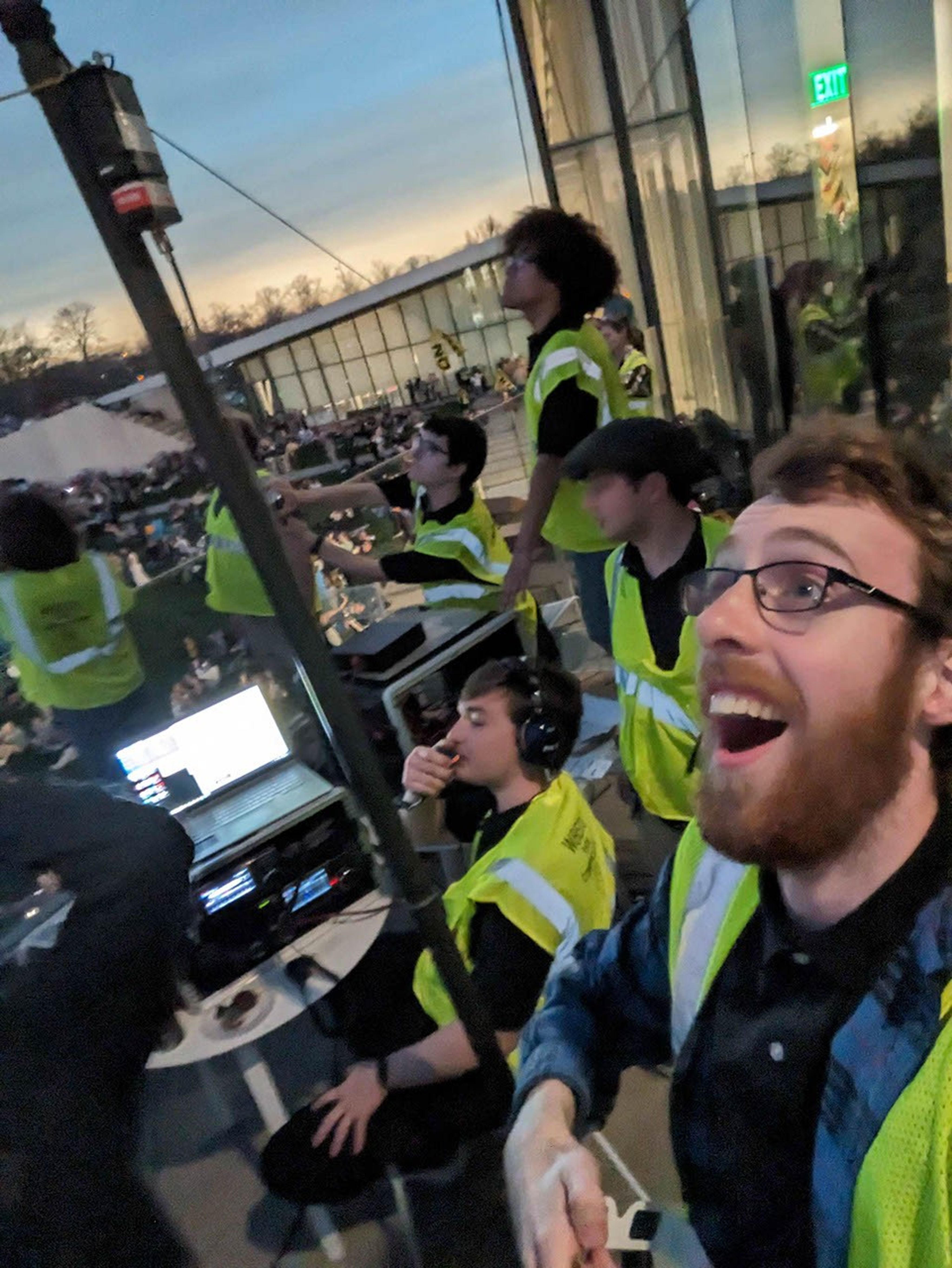 Several young people wearing yellow safety vests look upward. The lighting appears dusky, as it does during a total solar eclipse. One person is sitting at a table with radio equipment and a laptop with headphones on and a microphone in hand, while another person in the foreground looks up with a surprised but joyful expression.