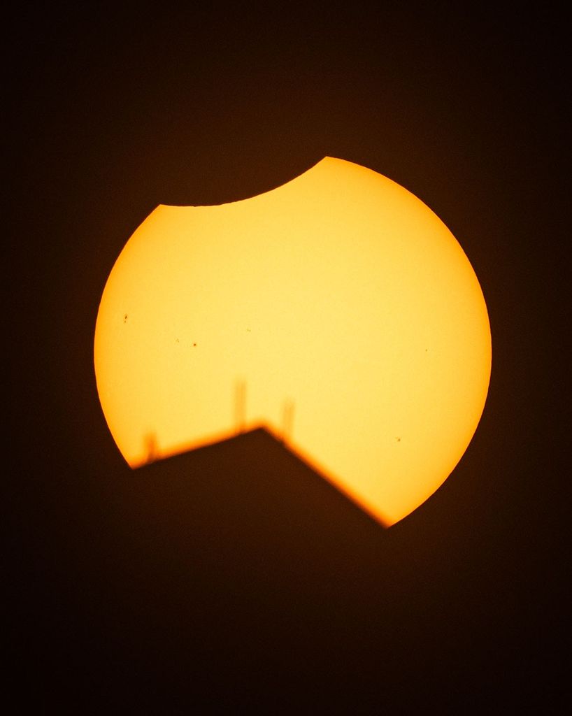 The Sun is shown partially eclipsed by the Moon, with a small, crescent-shaped piece apparently missing from the top portion of the Sun. A dark, triangular wedge also appears to be missing from the bottom of the Sun, caused by the top of a building blocking the Sun, with three antennas sticking up into the Sun from atop the building. A few sunspots can also be seen on the Sun on the left and in the lower right portions of the Sun.