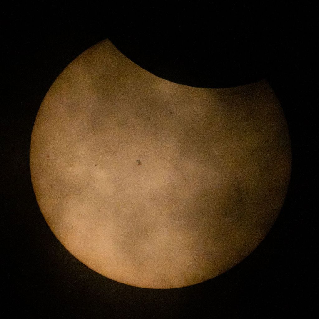 A partially eclipsed Sun appears with the Moon blocking a small portion of the Sun at top. Clouds also appear in front of the Sun. To the left of center is the small silhouette of the International Space Station. A few sunspots appear on the Sun to the left of the space station.