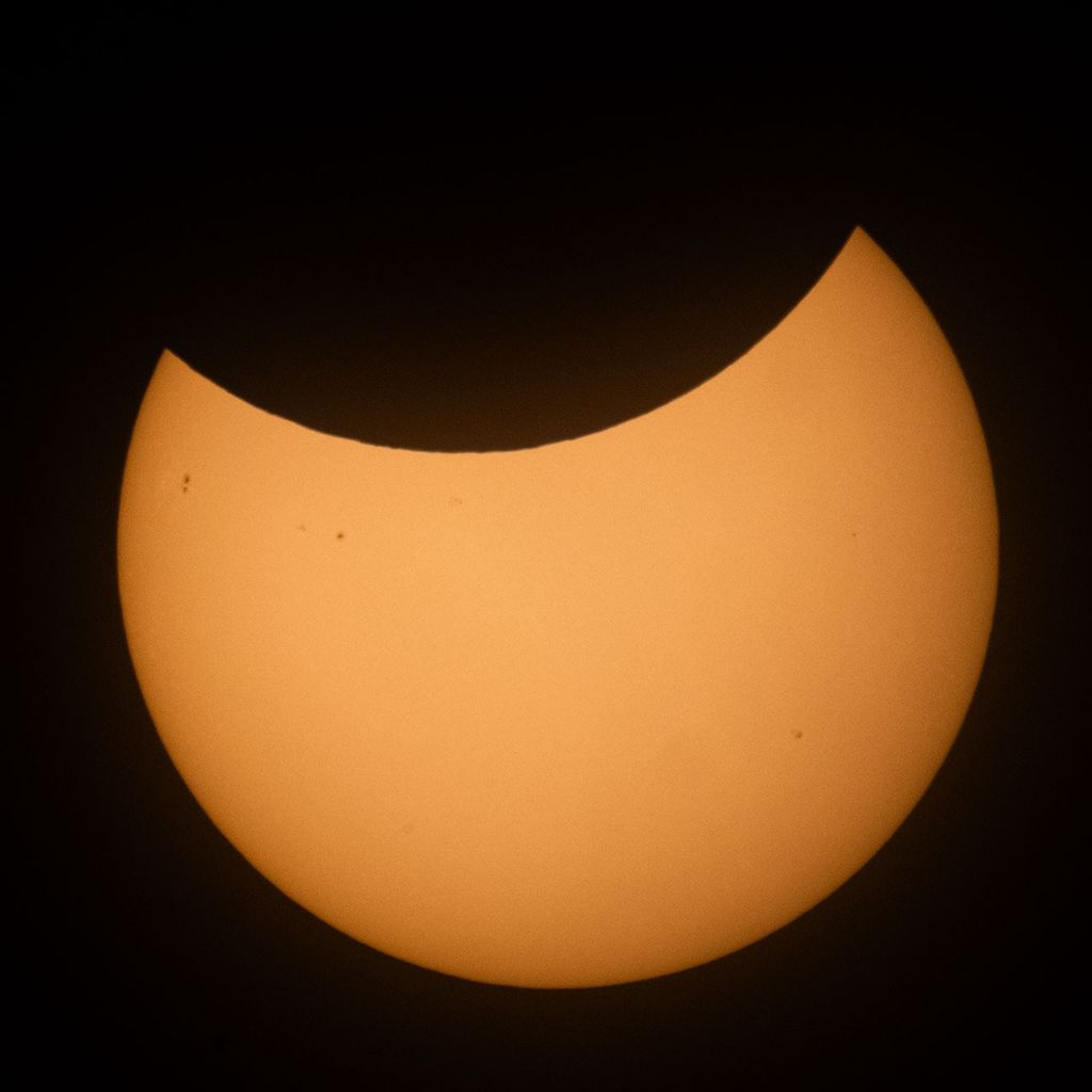 The Sun is shown partially eclipsed by the Moon, with a large, crescent-shaped piece apparently missing from the top portion of the Sun. A few sunspots can also be seen on the Sun on the left and in the lower right portions of the Sun.