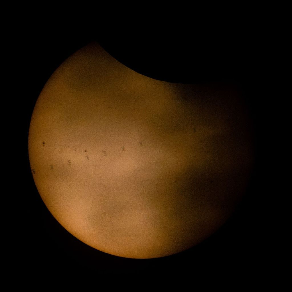 A partially eclipsed Sun appears with the Moon blocking a small portion of the Sun at top. Clouds also appear in front of the Sun. Crossing the Sun from the upper right to lower left is a series of silhouettes of the International Space Station. A few sunspots also appear on the left side of the Sun.