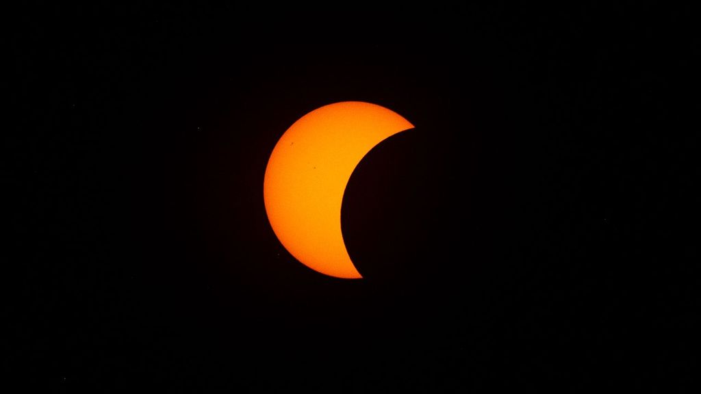A partially eclipsed Sun appears with the Moon blocking a little less than half of the Sun on the right side. A few sunspots appear in the upper left part of the Sun.