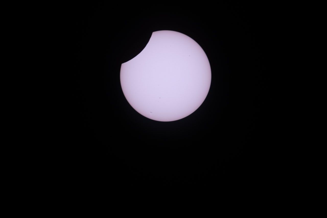 A photo shows the partially eclipsed Sun as a white disk, set against a black sky, with a small "bite" removed from the upper left portion of the solar disk.