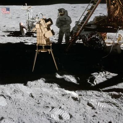An astronaut stands on the Moon near a lunar module and scientific equipment, with an American flag and lunar rover in the background. The lunar surface is covered in footprints and gray dust.