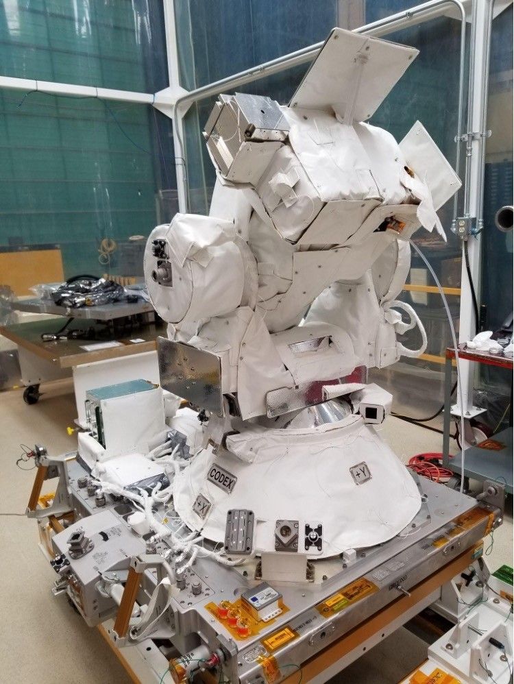 A large, white robotic instrument resembling a camera setup is mounted on a platform in a lab. It's enclosed in a transparent room, surrounded by various technical equipment and wires, with shelves and a cart nearby.