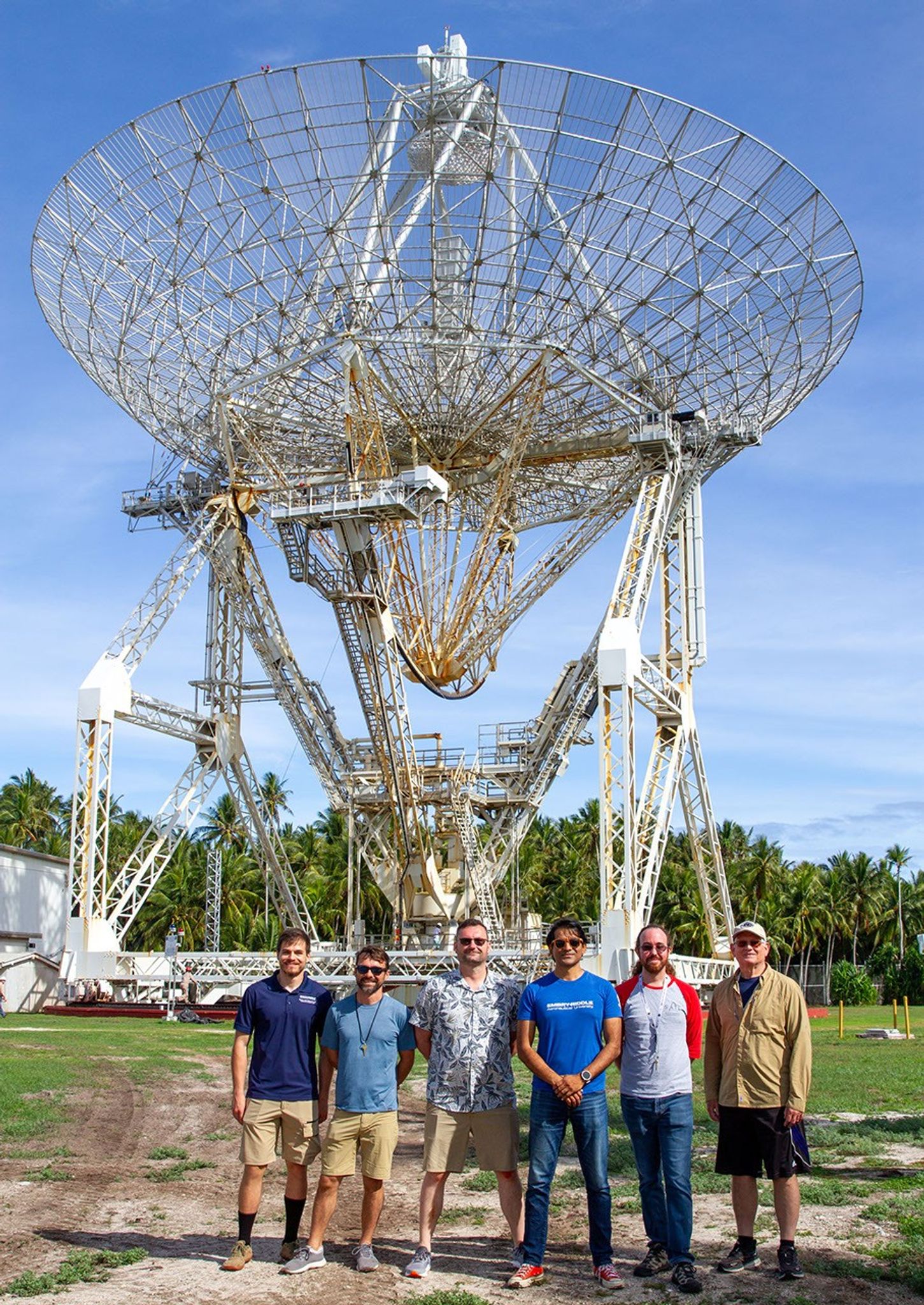 Six people stand in front of a large radio telescope dish under a blue sky, with palm trees in the background. The group is casually dressed and smiling, suggesting a scientific or research visit.