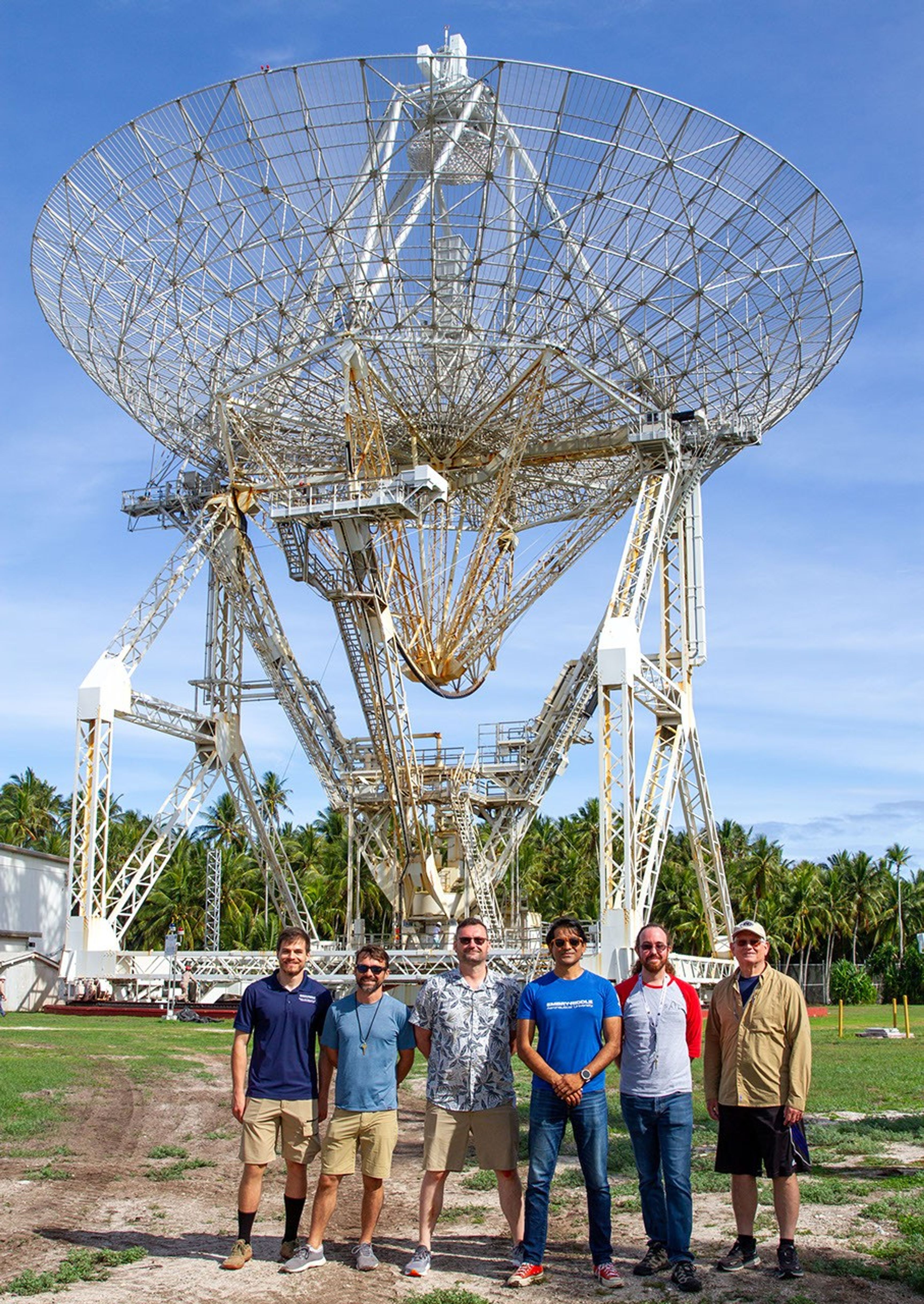 Six people stand in front of a large radio telescope dish under a blue sky, with palm trees in the background. The group is casually dressed and smiling, suggesting a scientific or research visit.