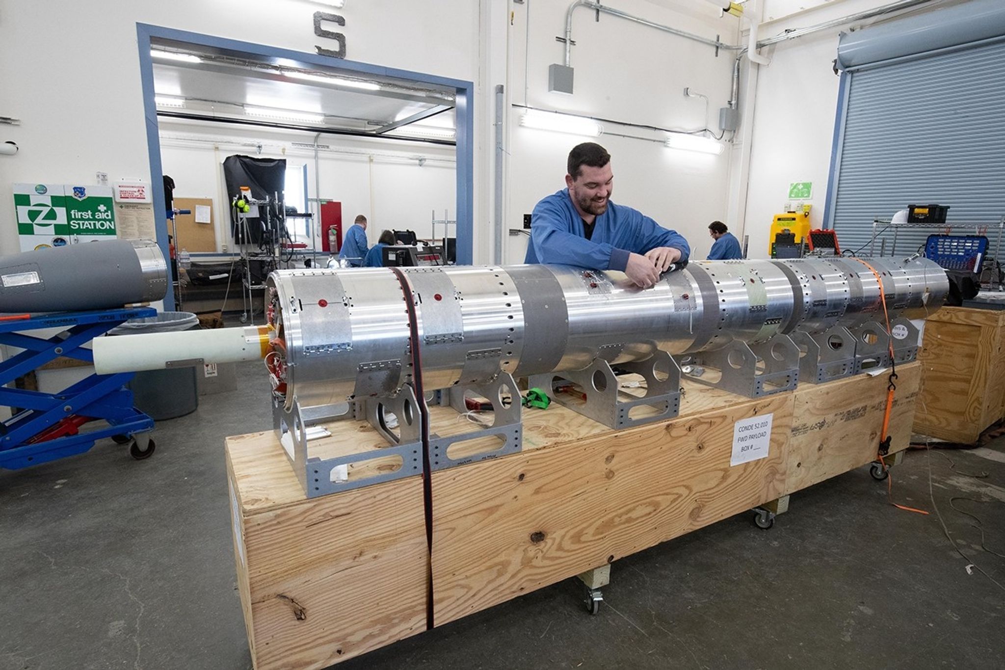 A technician in a blue static jacket leans over a long payload section of a sounding rocket laying horizontally. The payload is a silvery metal cylinder made up of many smaller cylinder sections put together. It sits upon a wooden box held up by many metal supports