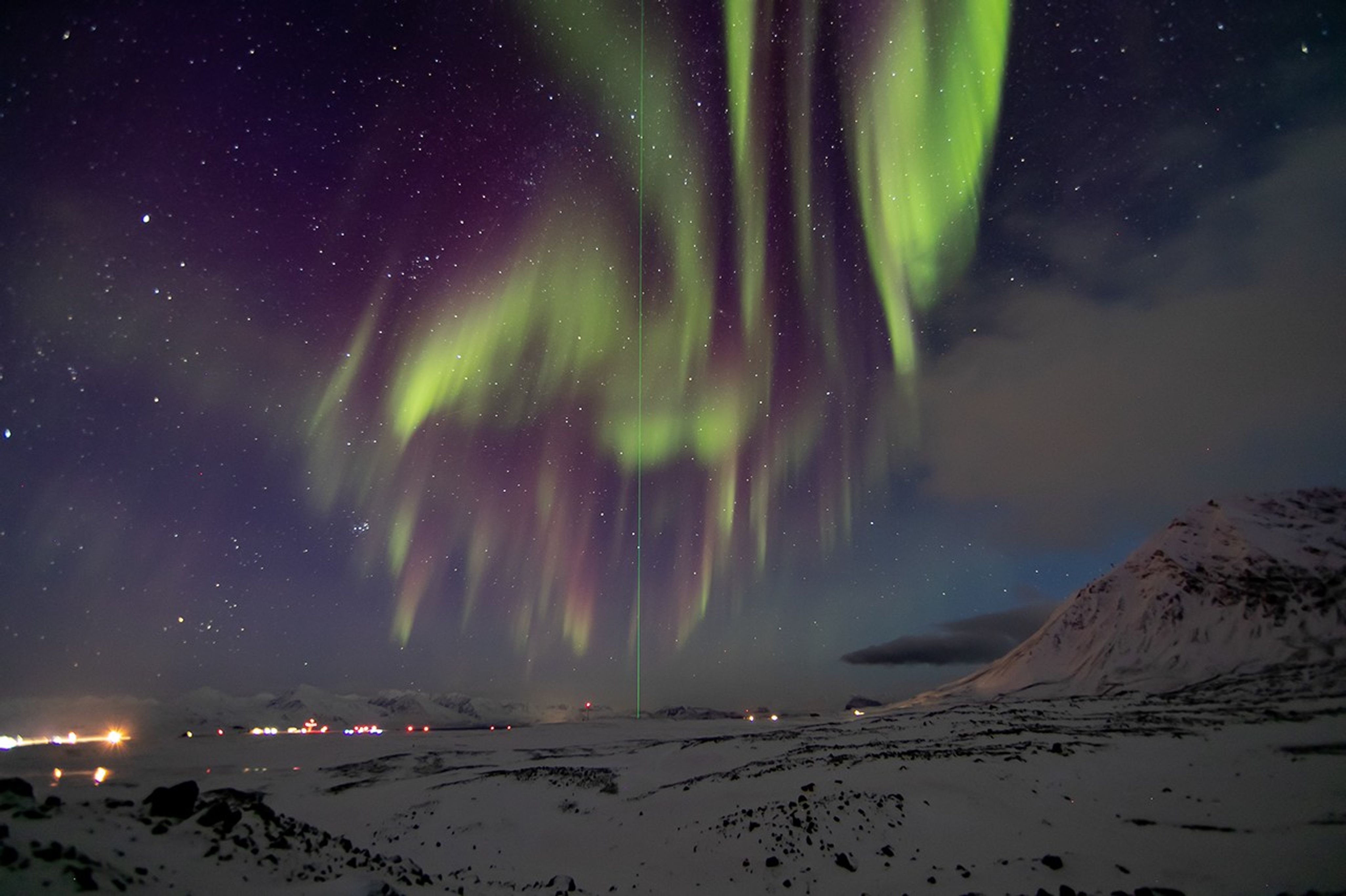 Bands of aurora fill part of a partially cloudy night sky. In the foreground, are snowy hills that lead to scattered lights in the distance. In the sky, the aurora appears as wavy bands — higher up, they appear purple and fade to green. In the middle of the image is a very thin, bright green vertical line that travels from the snowy landscape upward through the sky.