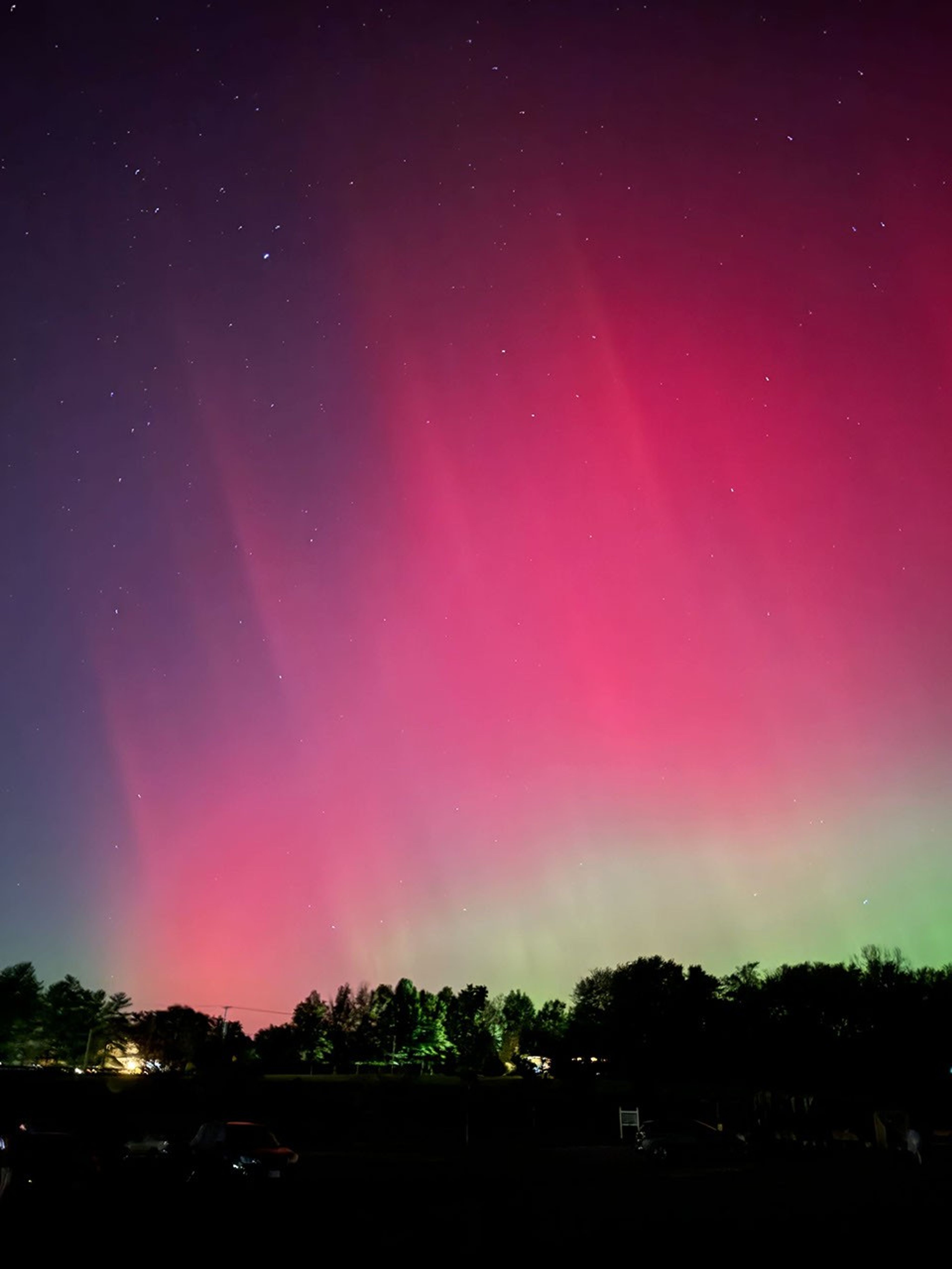 In the dark foreground, you care barely see cars and trees. Above that, huge swaths of pink aurora fill the sky. Closer to the trees the aurora turns green. Several stars are also scattered in the sky.