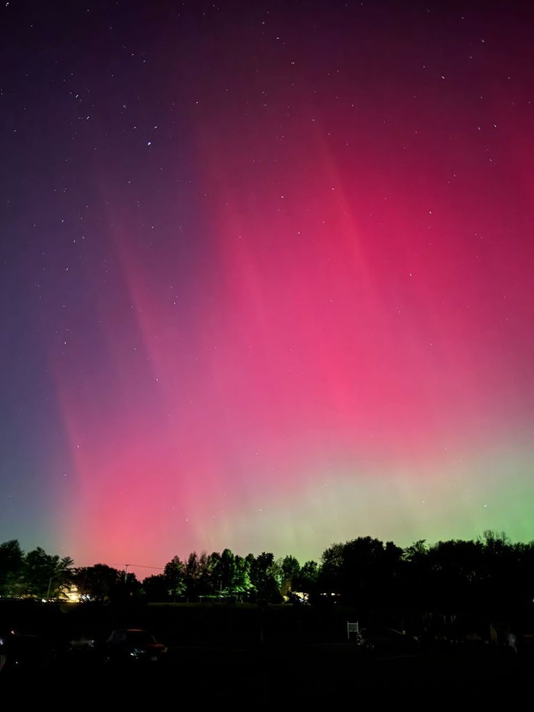 En el oscuro primer plano, apenas se ven coches y árboles. Por encima, enormes franjas de aurora rosa llenan el cielo. Más cerca de los árboles, la aurora se vuelve verde. Varias estrellas también se esparcen por el cielo.