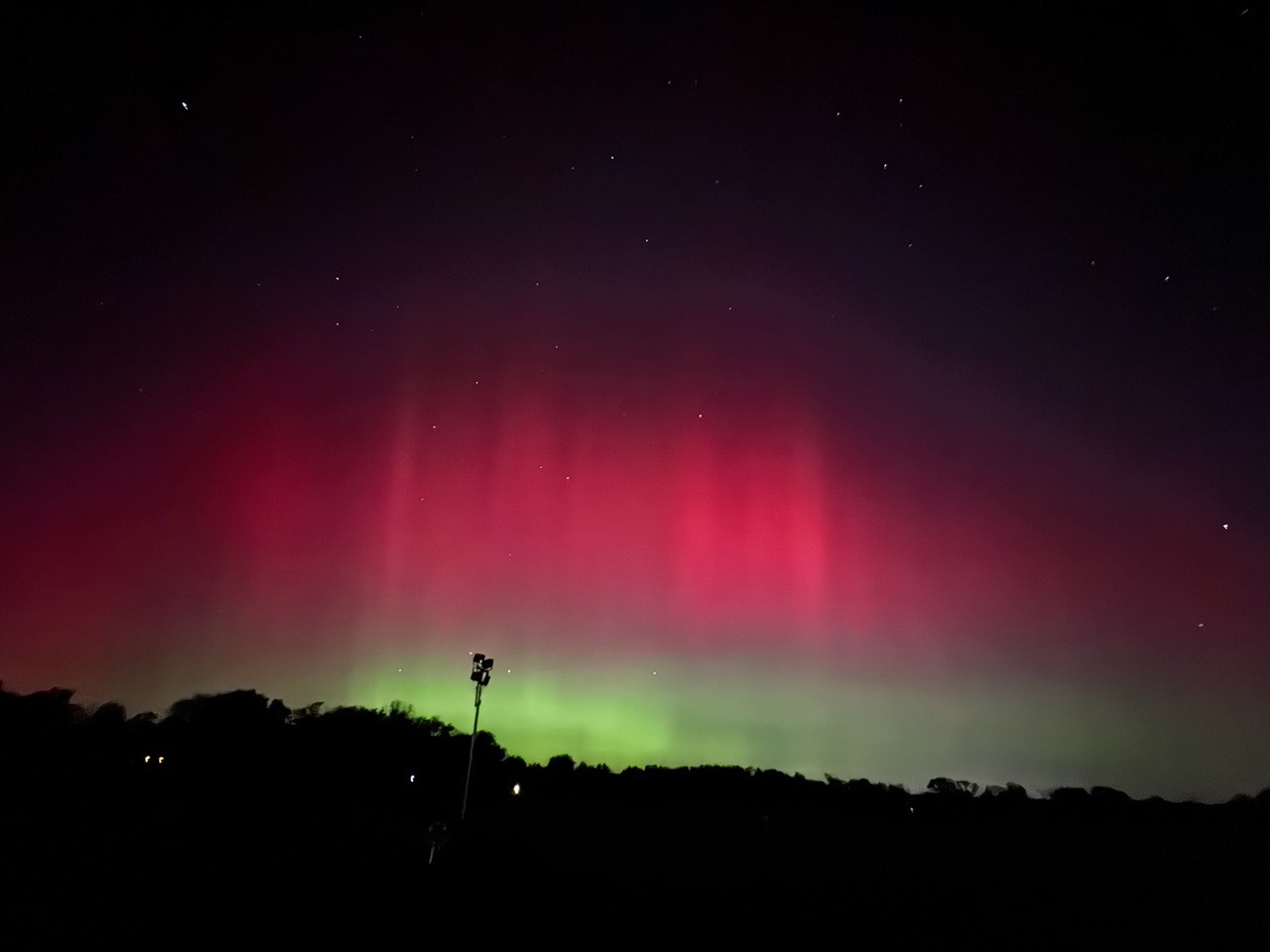In the foreground, is a very dark landscape with trees and one tall light post. Above the landscape is a large swatch of aurora that fills the middle of the image horizontally. It is bright green toward the bottom and bright pink toward the top. Fill the top of the image is the night sky with some stars.