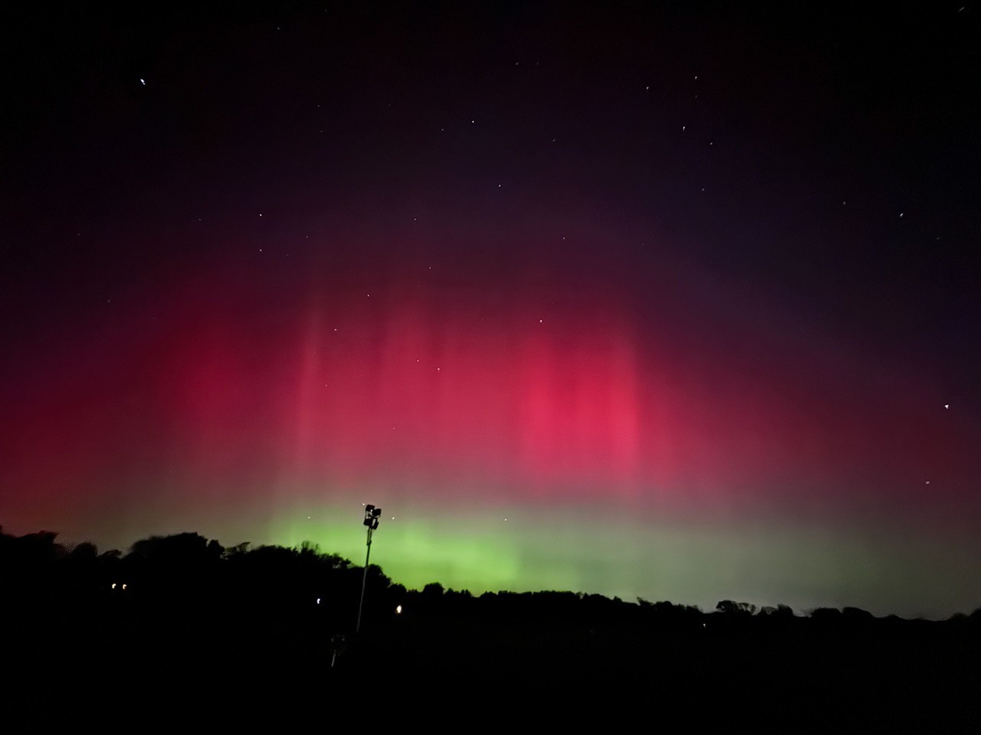 In the foreground, is a very dark landscape with trees and one tall light post. Above the landscape is a large swatch of aurora that fills the middle of the image horizontally. It is bright green toward the bottom and bright pink toward the top. Fill the top of the image is the night sky with some stars.