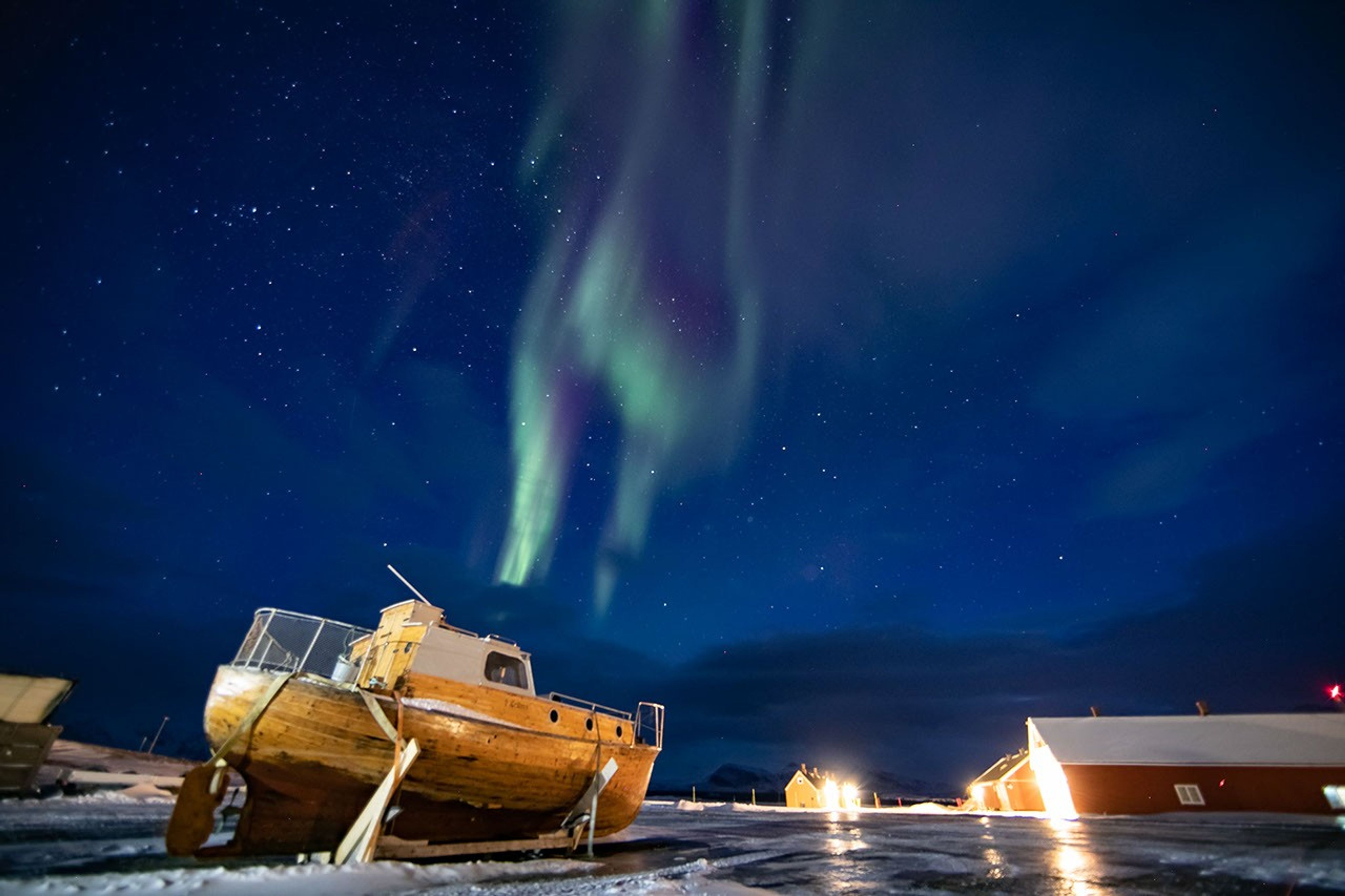 In the foreground is a yellow boat, stuck in snow in ice. There are also small buildings. In the sky, which appears dark blue with stars, are bands of green and purple aurora.