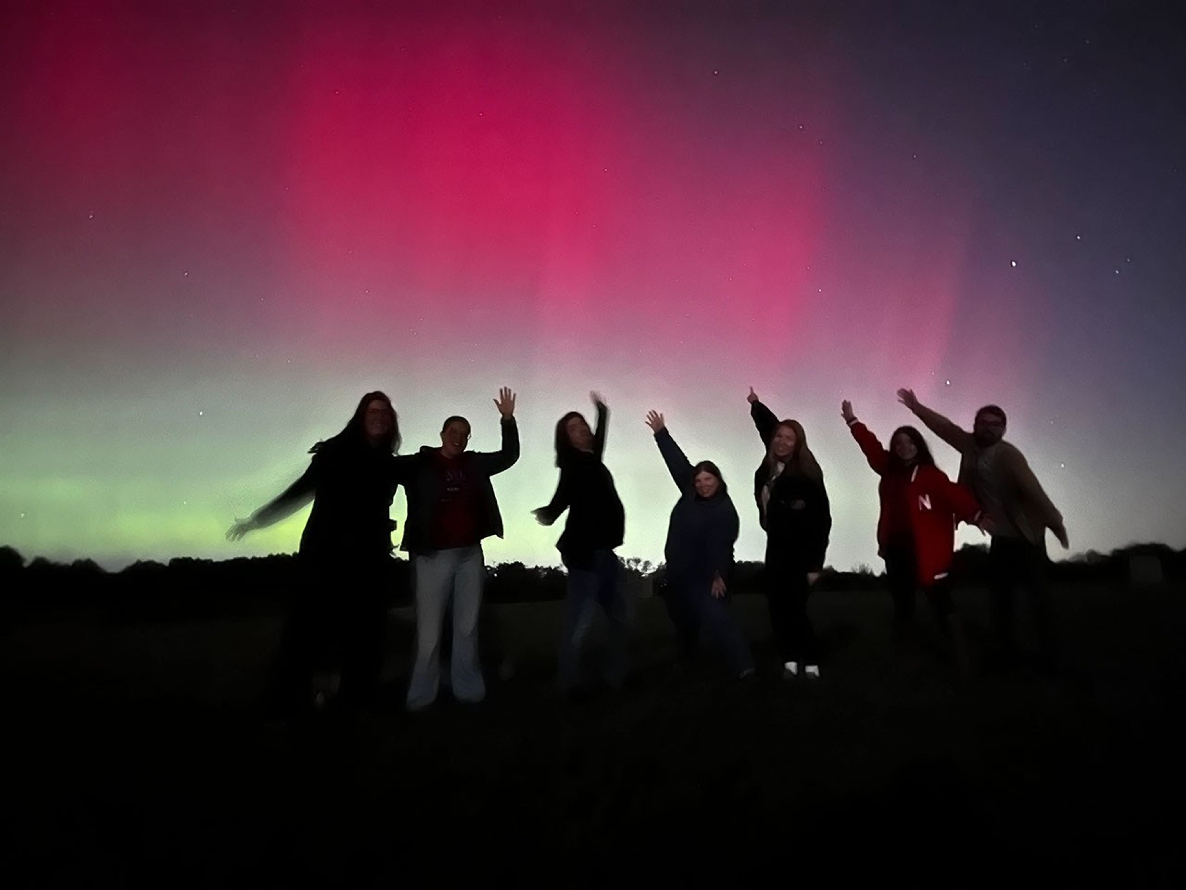 A group of seven people stand in front of an aurora. They are jumping with their hands in the air. Behind them, the sky is mostly filled with the pink and green of an aurora.