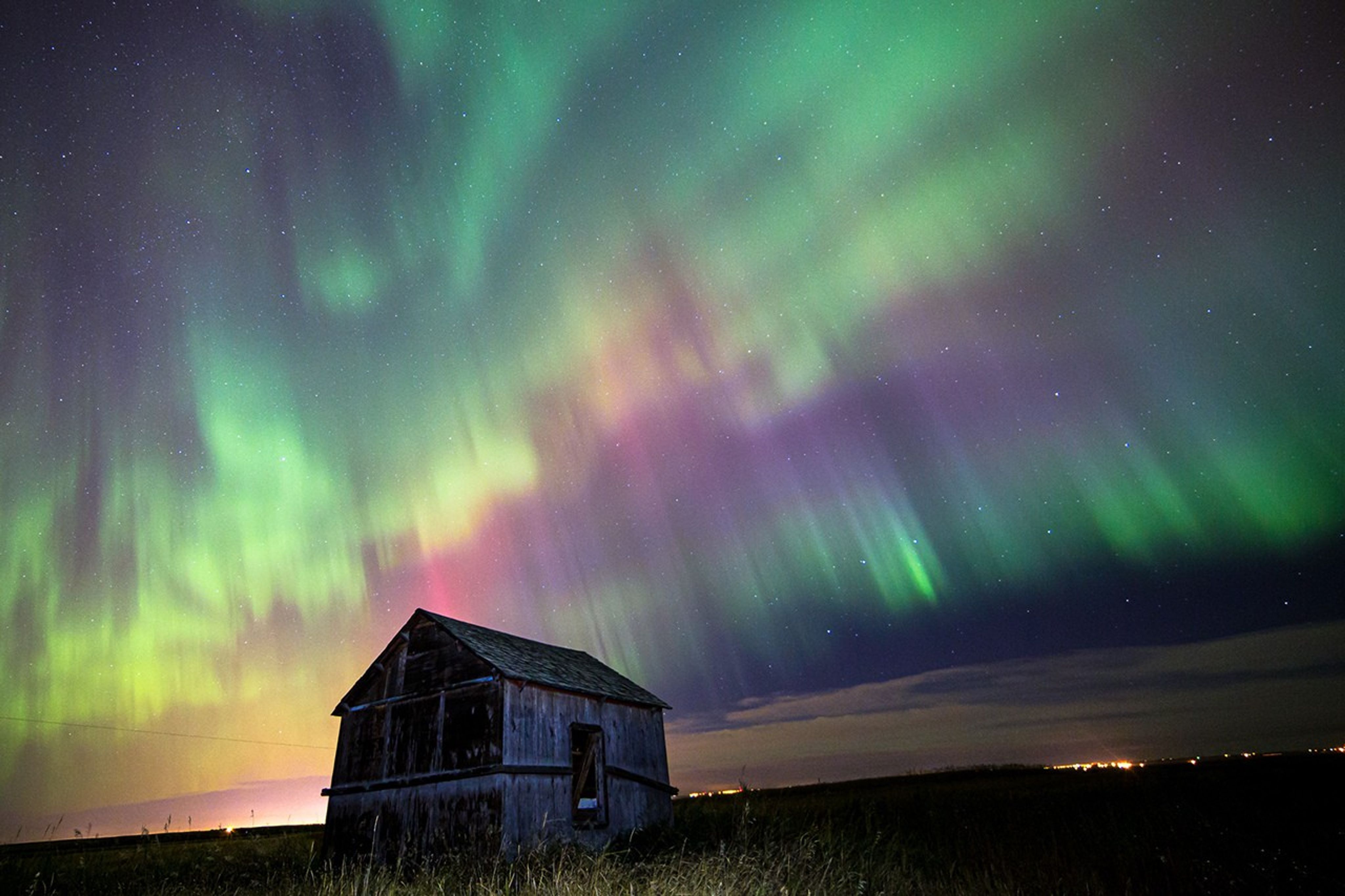 There is a house, alone in an empty grassy field at night. Above the house are bands of green, purple, and pink aurora. In the distance, some lights can be seen.