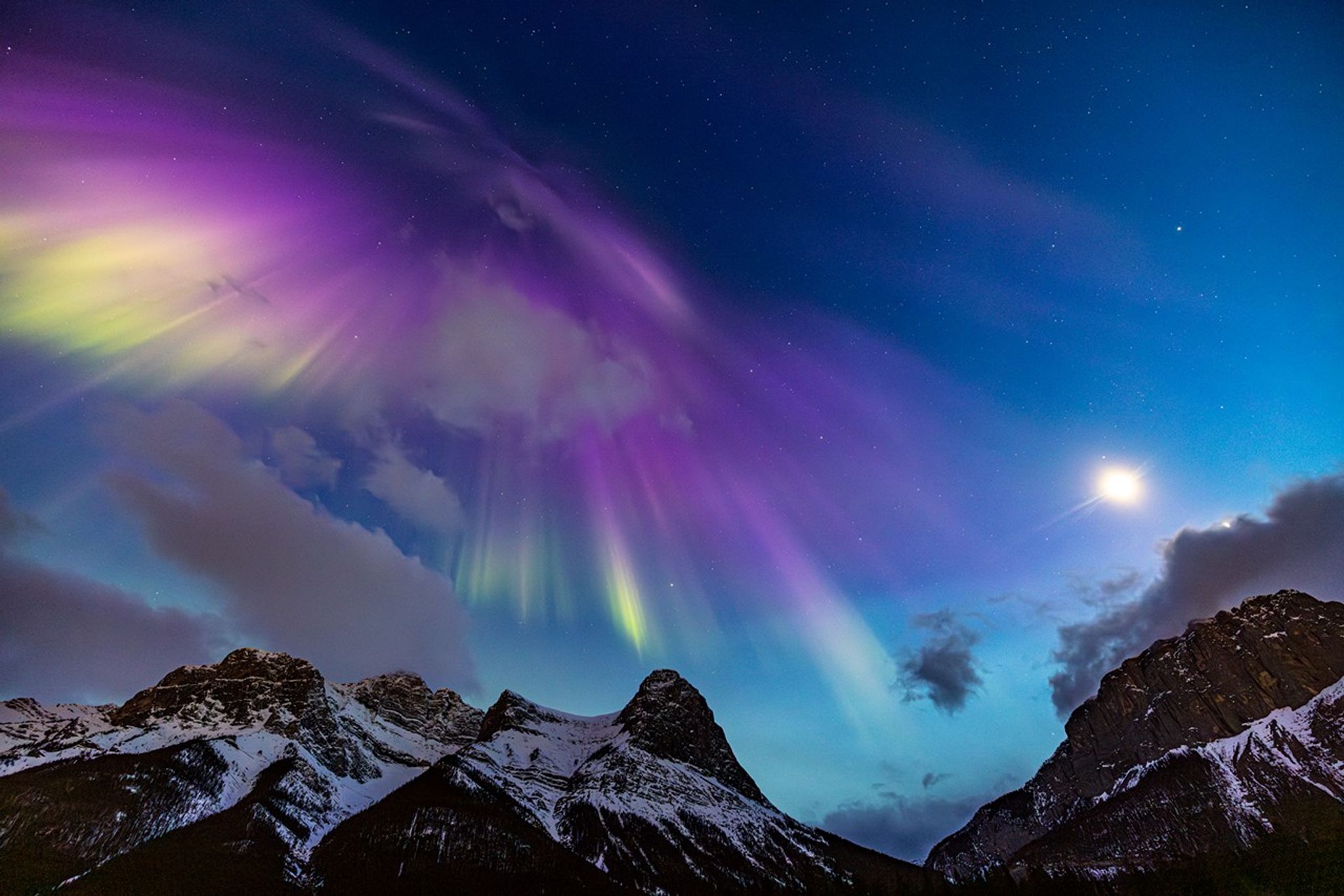 In the foreground are snowcapped mountains. Against a blue sky, a bright purple, yellow, and green aurora fills most of the sky. Some clouds are scattered as well.