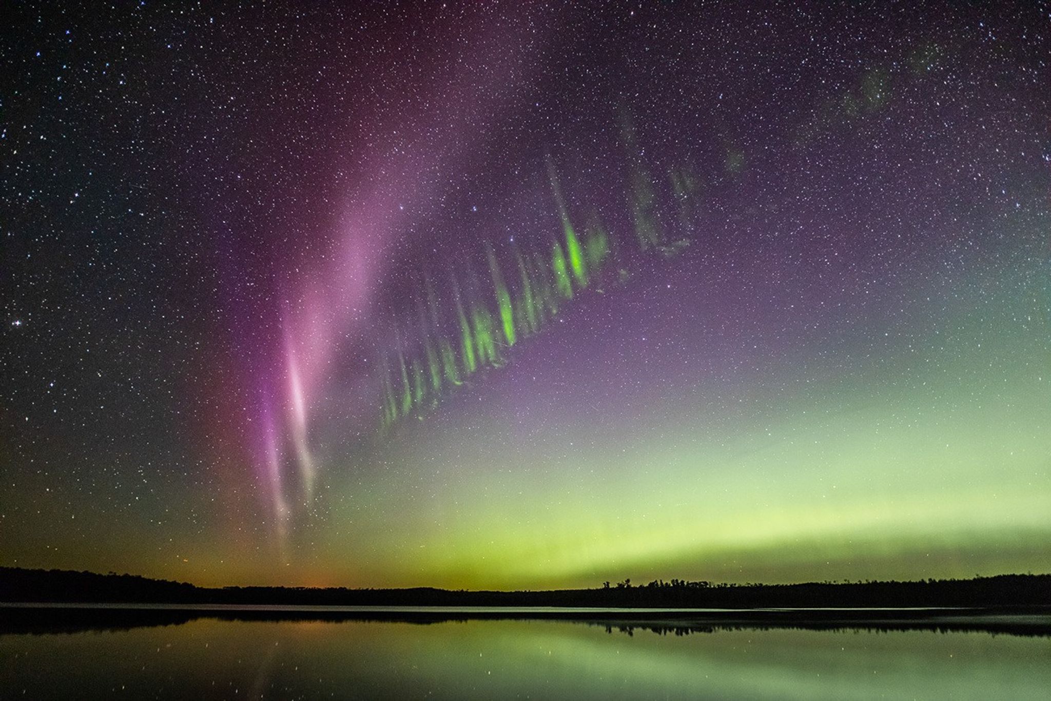 In the foreground is a flat body of water. Green aurora and stars are reflected on the water's surface. In the sky is a purple band of aurora traveling diagonally across the image from lower left to upper right. Parallel and below to it are vertical stripes of green.