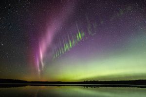 In the foreground is a flat body of water. Green aurora and stars are reflected on the water's surface. In the sky is a purple band of aurora traveling diagonally across the image from lower left to upper right. Parallel and below to it are vertical stripes of green.