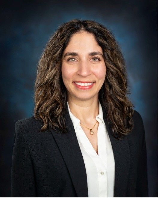 Photo of a smiling woman with dark, wavy hair, white blouse, and dark blazer