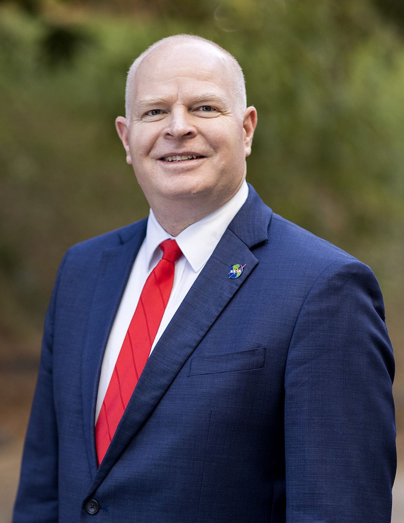 Photo of a smiling man in a blue suit, white shirt, and red tie