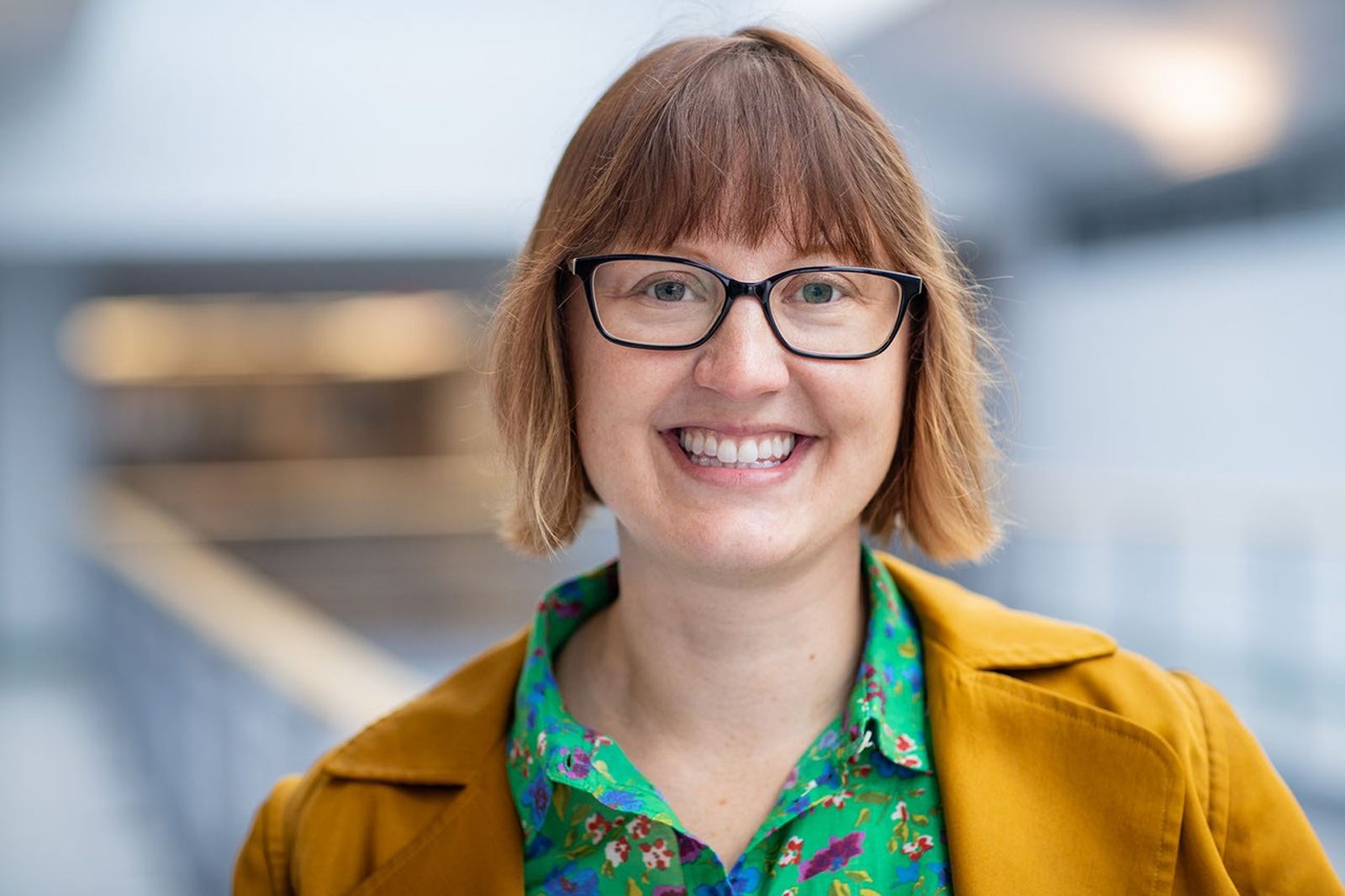 Photo of a smiling woman with glasses, a green print shirt and yellow blazer