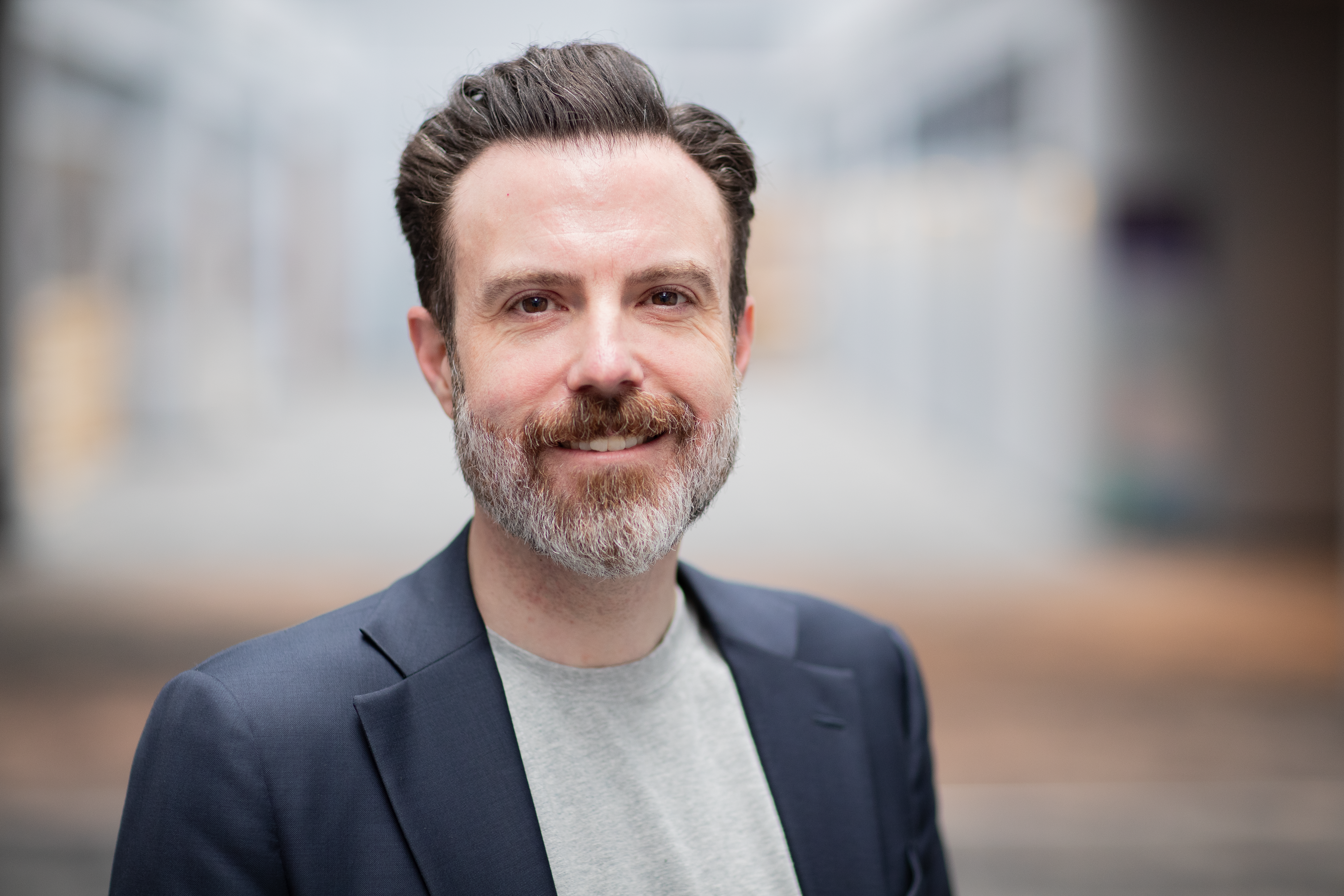 Portrait of a smiling man with a beard wearing a grey shirt and dark blazer.