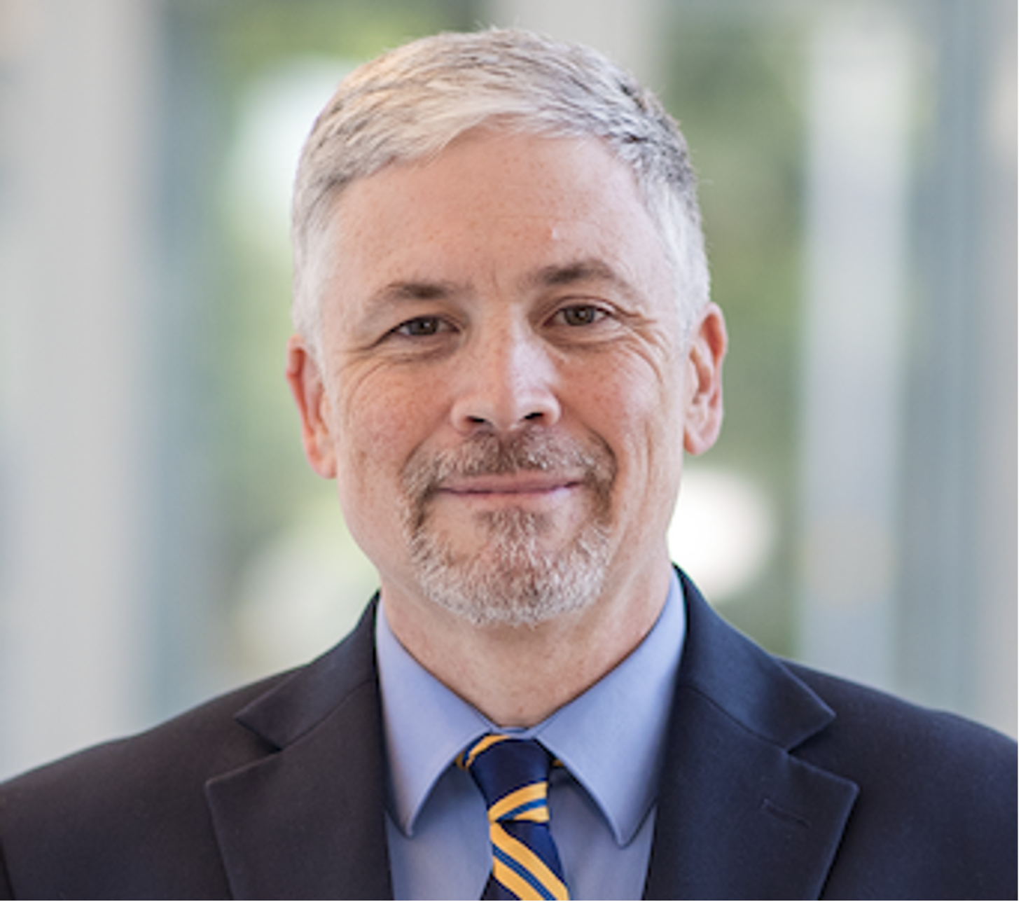 Photo of a smiling man with dark blue blazer, light blue shirt, and blue and yellow striped tie.