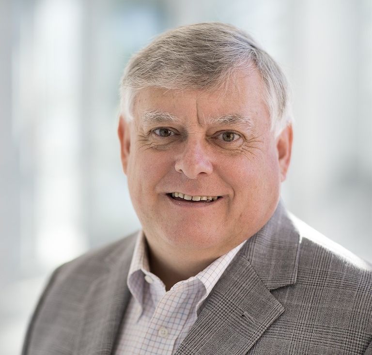Photo of a smiling man standing in front of a window wearing a checkered shirt and plaid blazer.