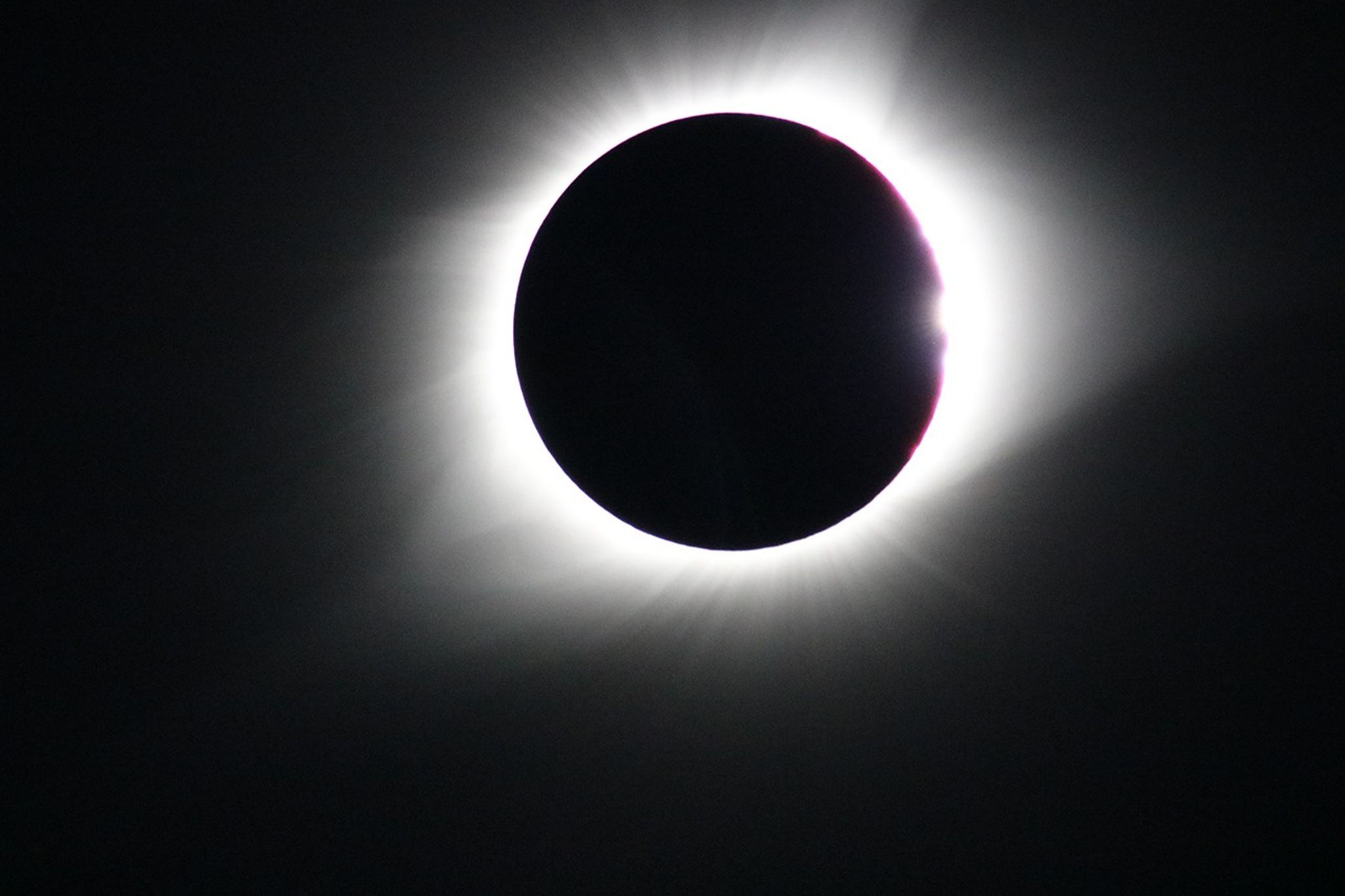 A total solar eclipse showing the dark silhouette of the Moon completely covering the Sun, with the Sun’s bright corona radiating around the edges against a dark sky. A faint sliver of red appears on the upper right of the eclipse, showing the Sun's chromosphere.