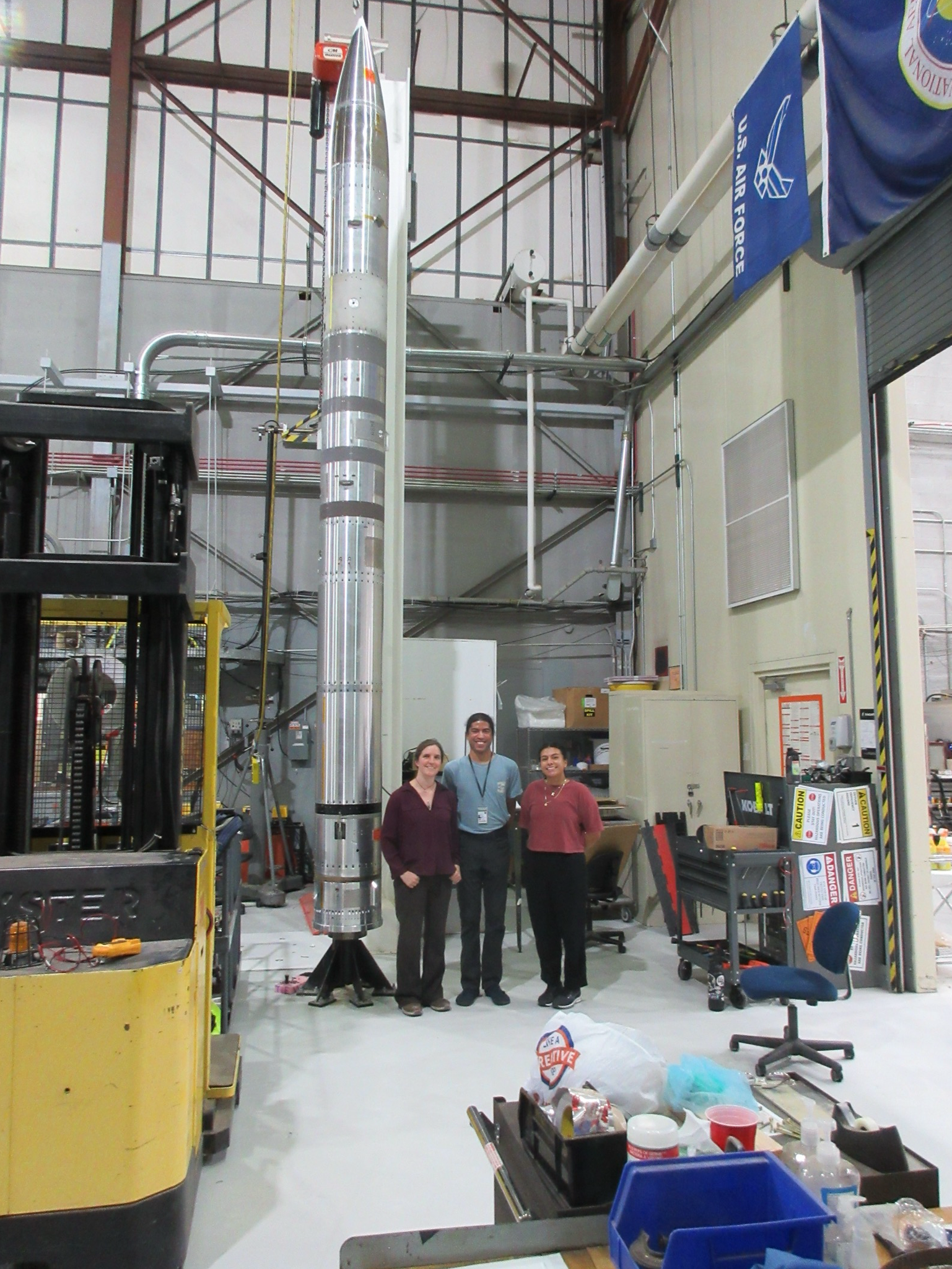 Three people stand smiling in front of a tall, silver rocket inside an industrial workshop. The workspace contains equipment, tools, a forklift, and an Air Force flag hanging above.