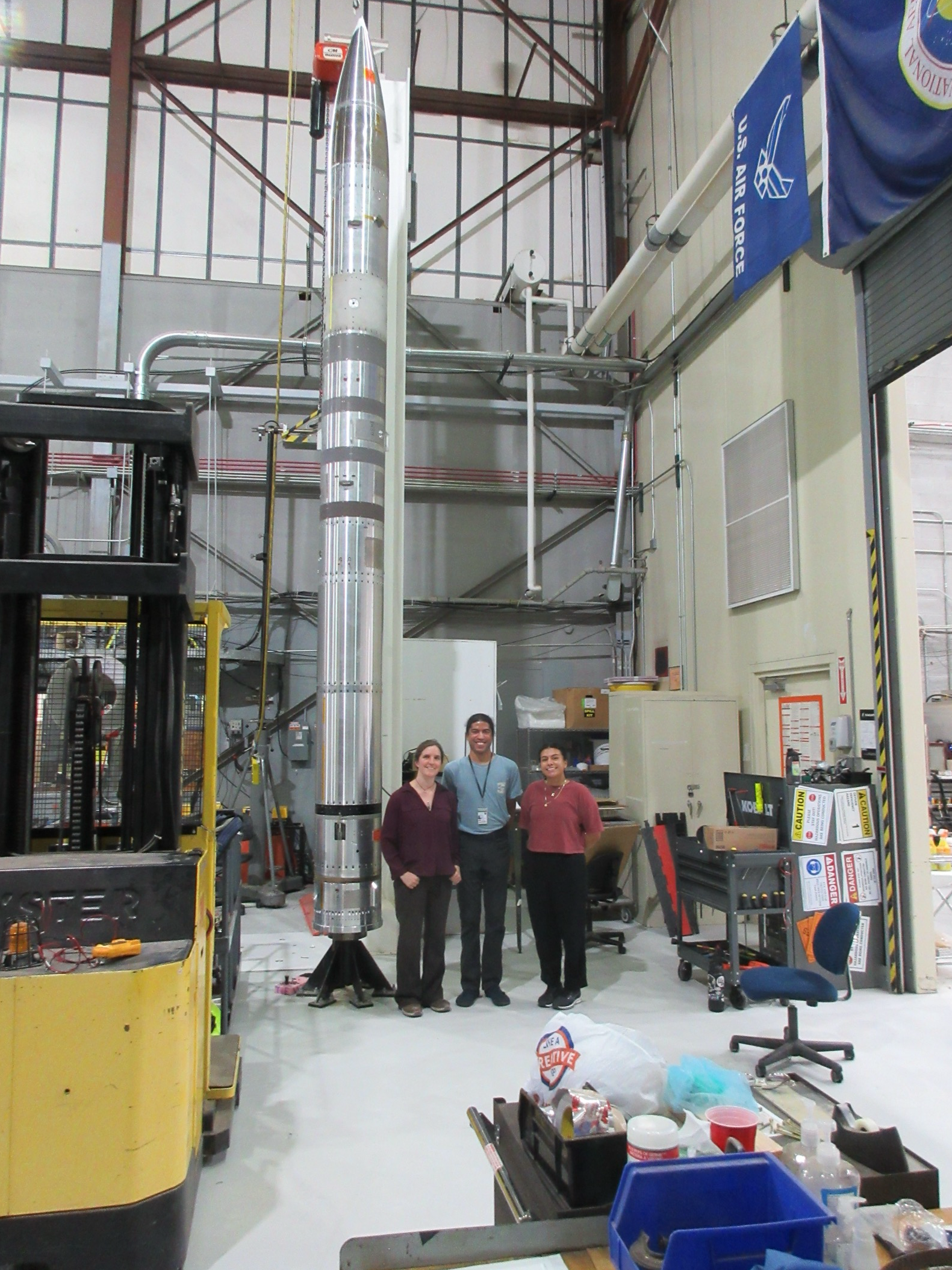 Three people stand smiling in front of a tall, silver rocket inside an industrial workshop. The workspace contains equipment, tools, a forklift, and an Air Force flag hanging above.